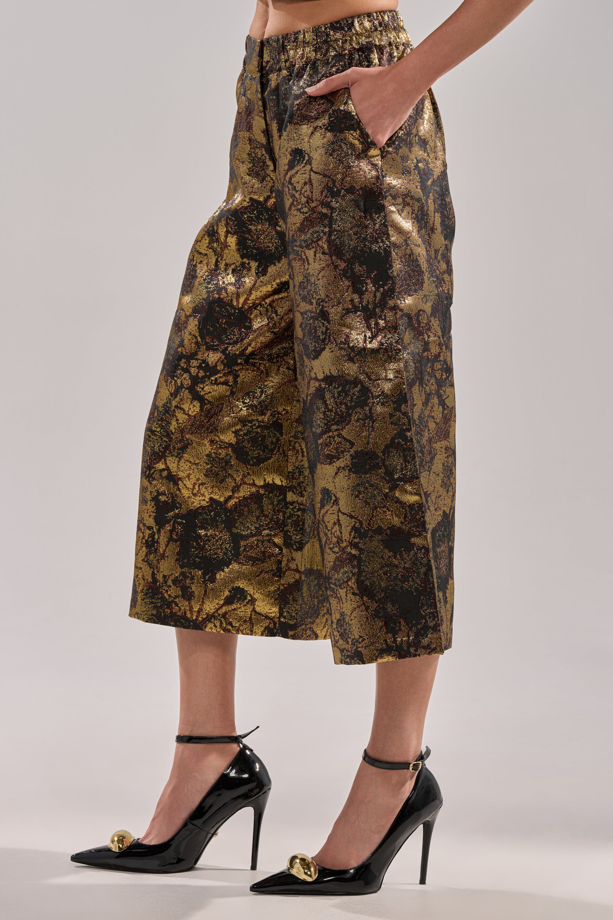 A person wearing the LOTUS BROCADE CAPRI in gold and black with wide-leg cropped design, hands in pockets, paired with black high heels featuring gold decorative accents on the toes.