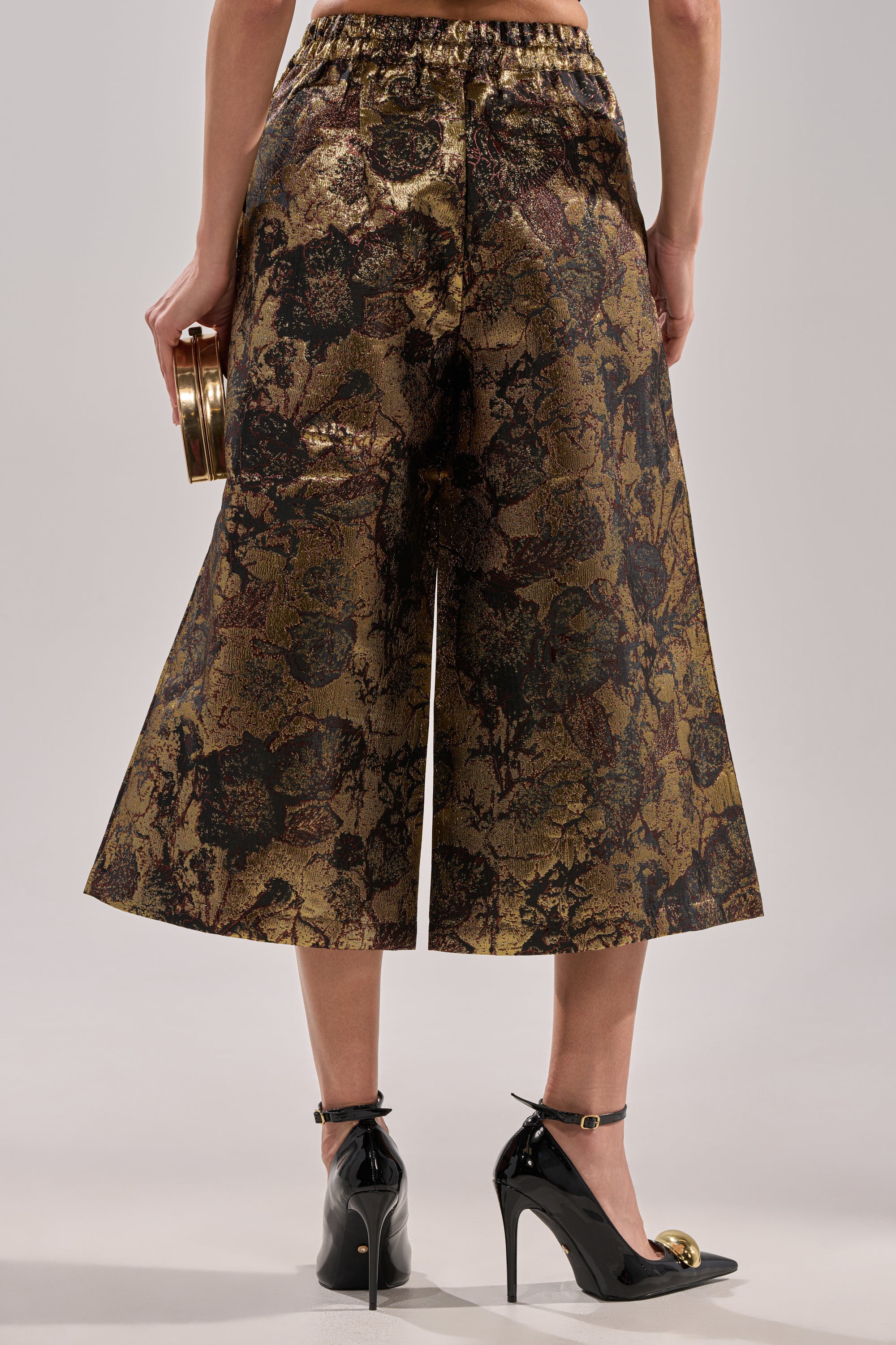 A person wearing LOTUS BROCADE CAPRI wide-leg cropped pants in metallic gold and black, paired with black ankle-strap heels and a small round gold clutch, is shown from the back against a neutral background.