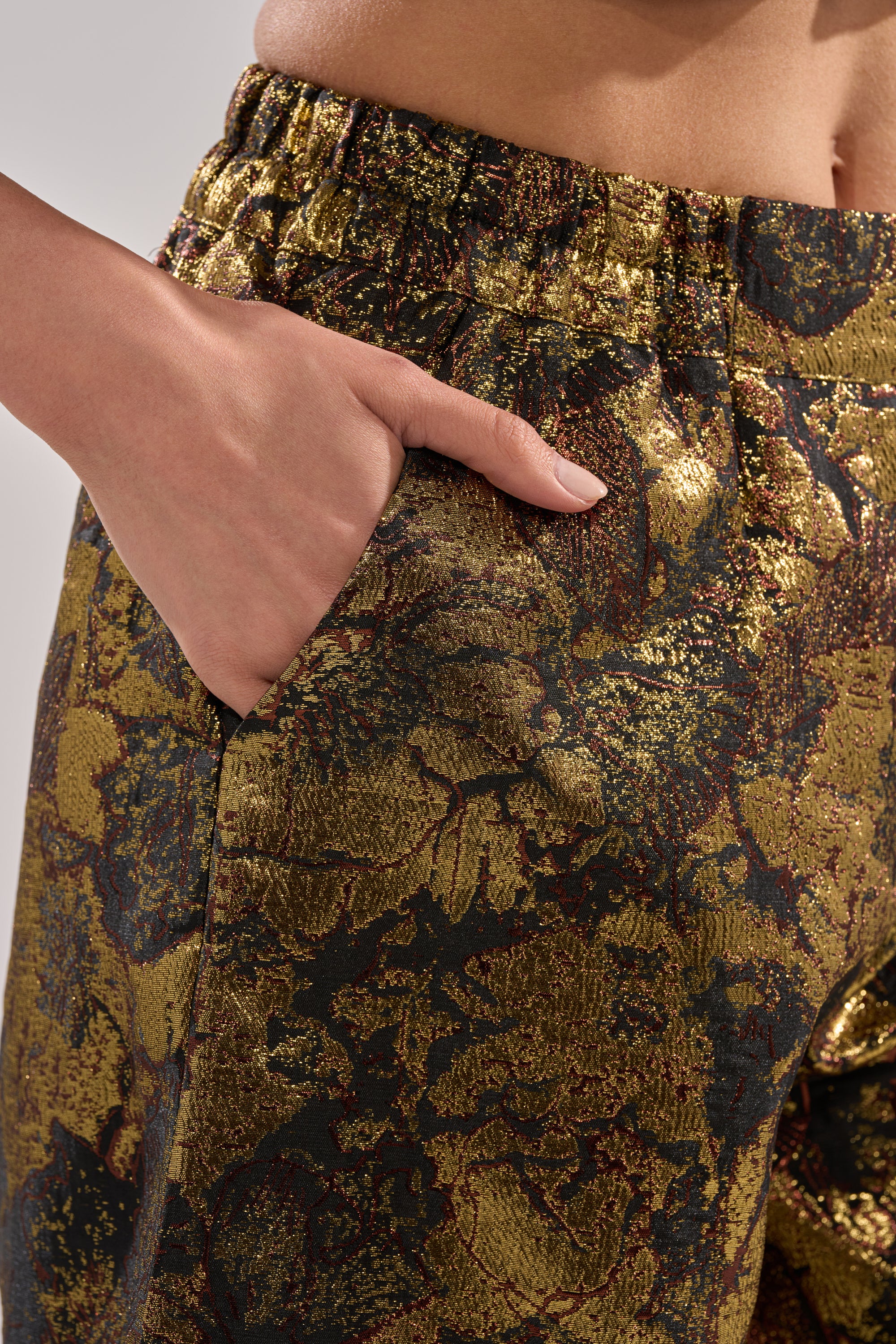 A close-up shows a person wearing the LOTUS BROCADE CAPRI in metallic gold with a dark floral pattern. Their right hand is in the pocket, drawing attention to the capri’s unique textured fabric and stylish design.