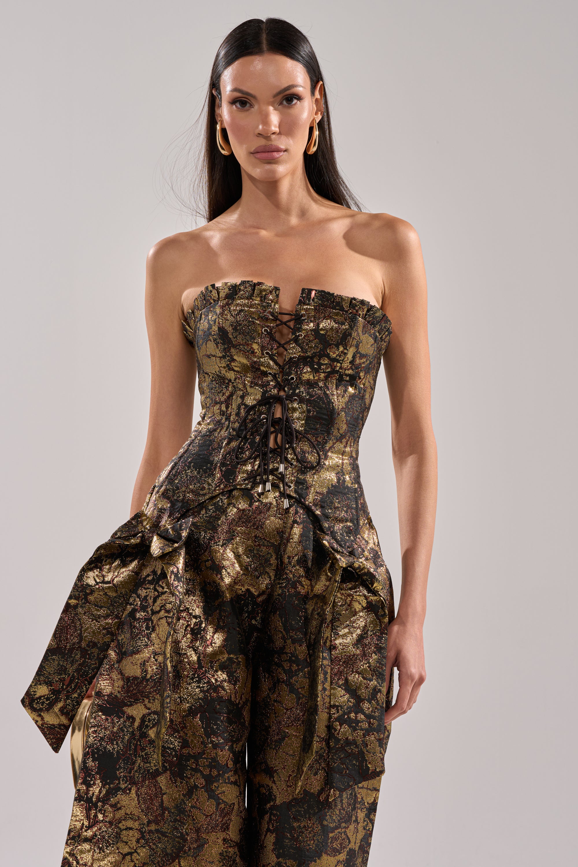 A woman with long dark hair wears the LOTUS BROCADE CORSET, a gold-and-black floral strapless top with a lace-up front, paired with matching high-waisted wide-leg pants against a plain light background.