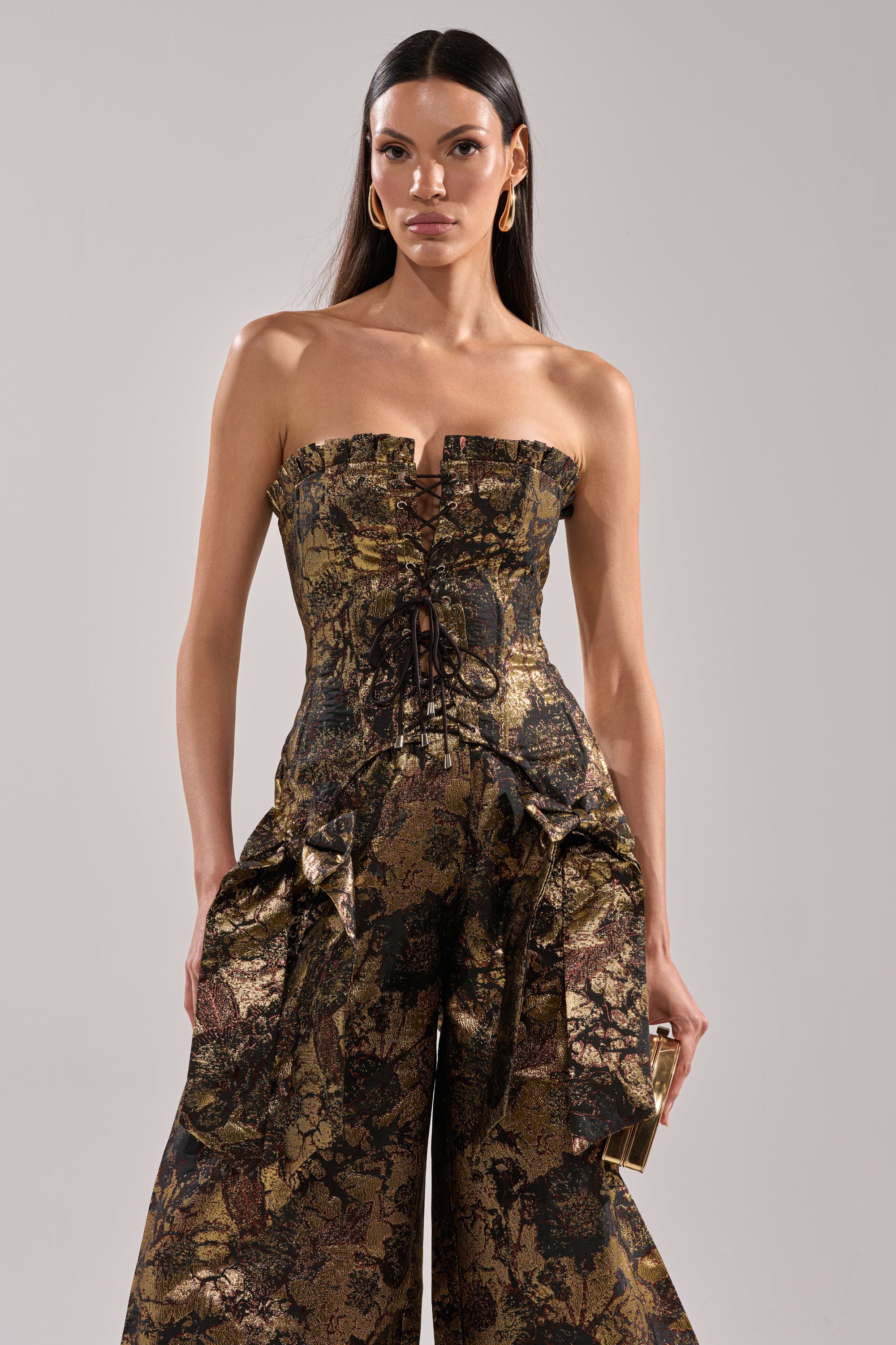 A woman in the LOTUS BROCADE CORSET, a strapless metallic jumpsuit with lace-up front and wide-leg pants, stands against a plain light background, holding a gold clutch.