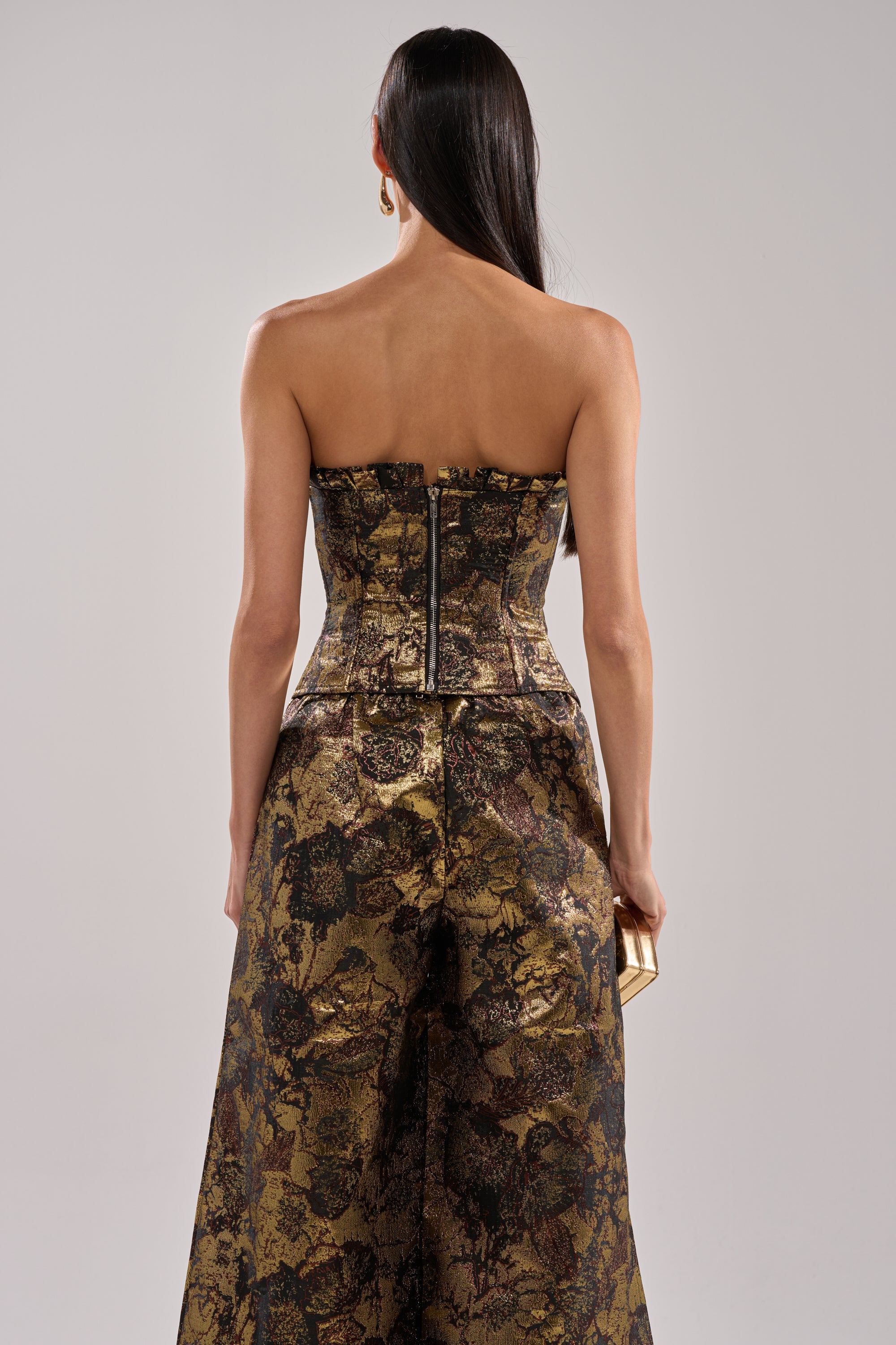 A woman with long dark hair stands with her back to the camera against a light gray background, wearing the LOTUS BROCADE CORSET in gold-and-black floral brocade and matching wide-leg pants, holding a small gold clutch.