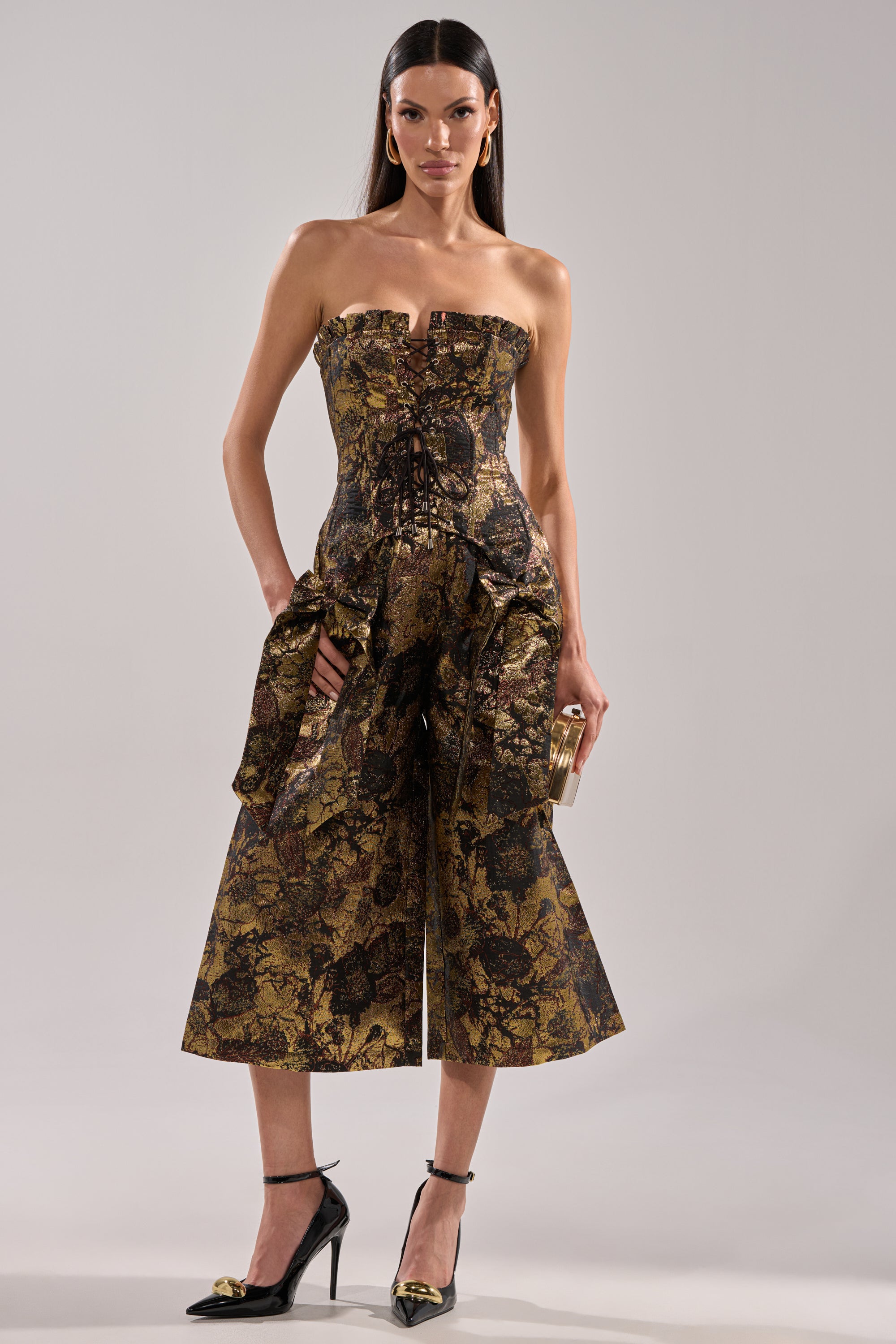 A woman faces forward in the LOTUS BROCADE CORSET, a gold-and-black floral corset jumpsuit featuring a lace-up front and flared culotte pants. She accessorizes with a small clutch and black ankle-strap heels.