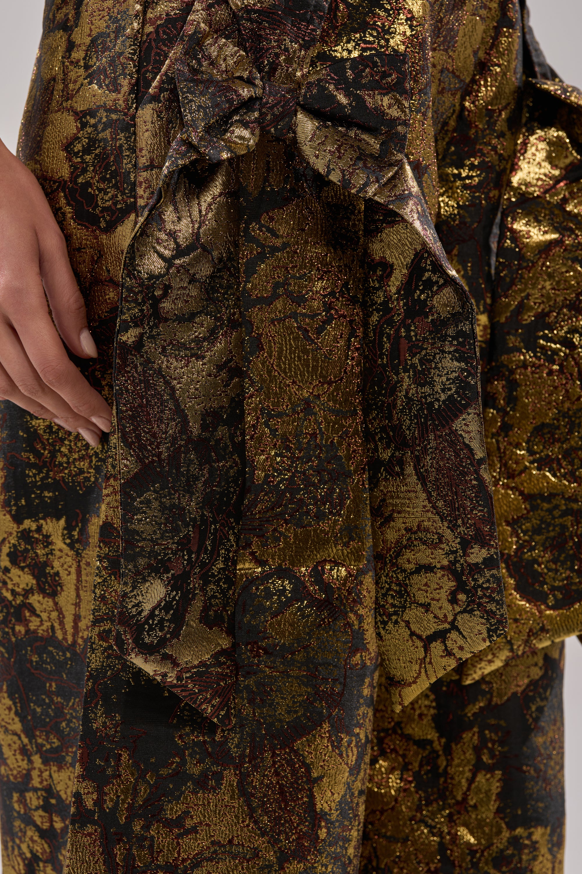 Close-up of a hand resting on shiny, textured fabric in gold, black, and brown floral patterns—a detail from the LOTUS BROCADE CORSET with elegant folds and a decorative bow.