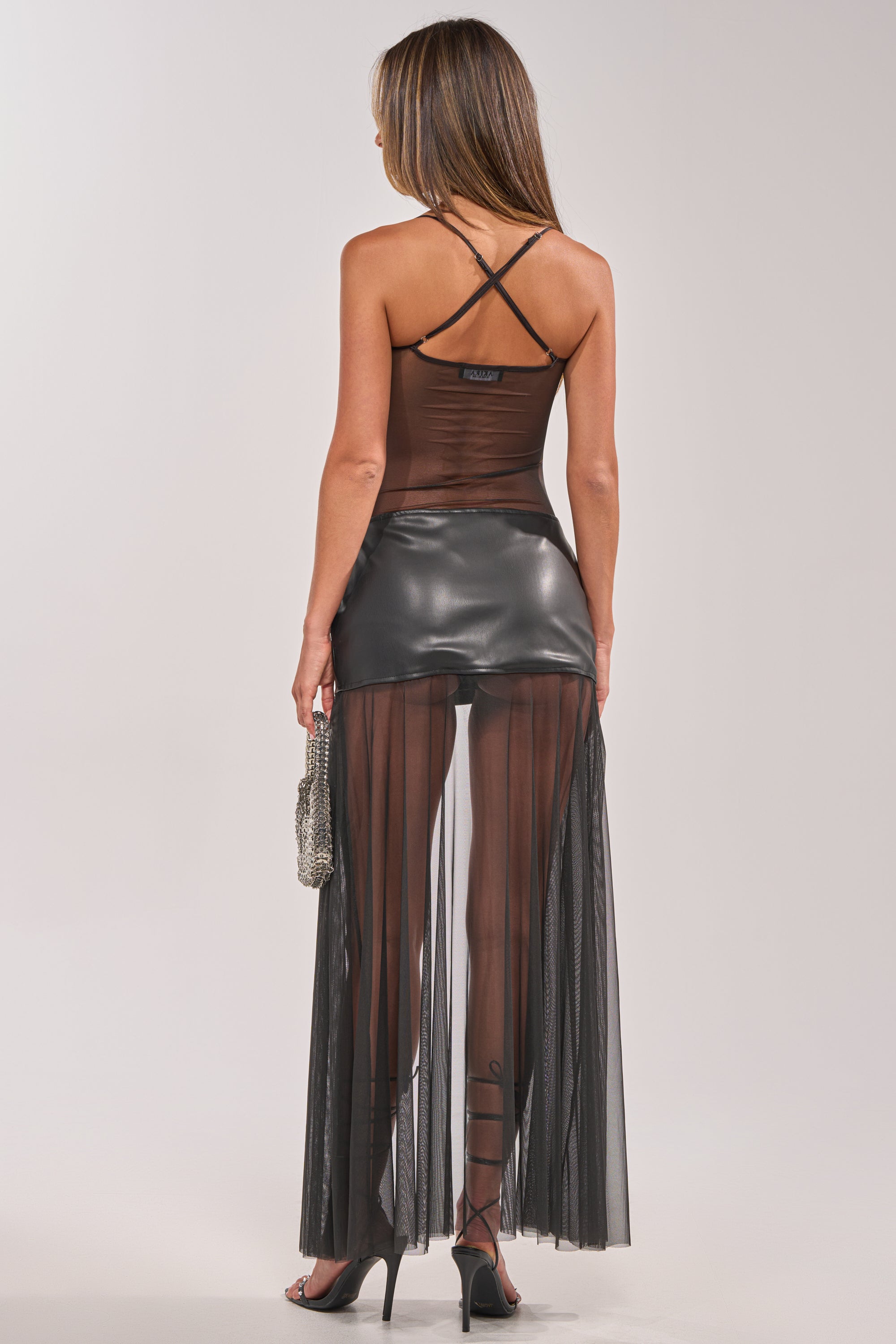 Woman with her back to the camera wears the TRUE ICON CUT OUT MINI DRESS, featuring mesh panels, a crisscross strap top, and a shiny black mini skirt. She carries a handbag and pairs the look with black high heels—an ideal statement evening outfit. 3