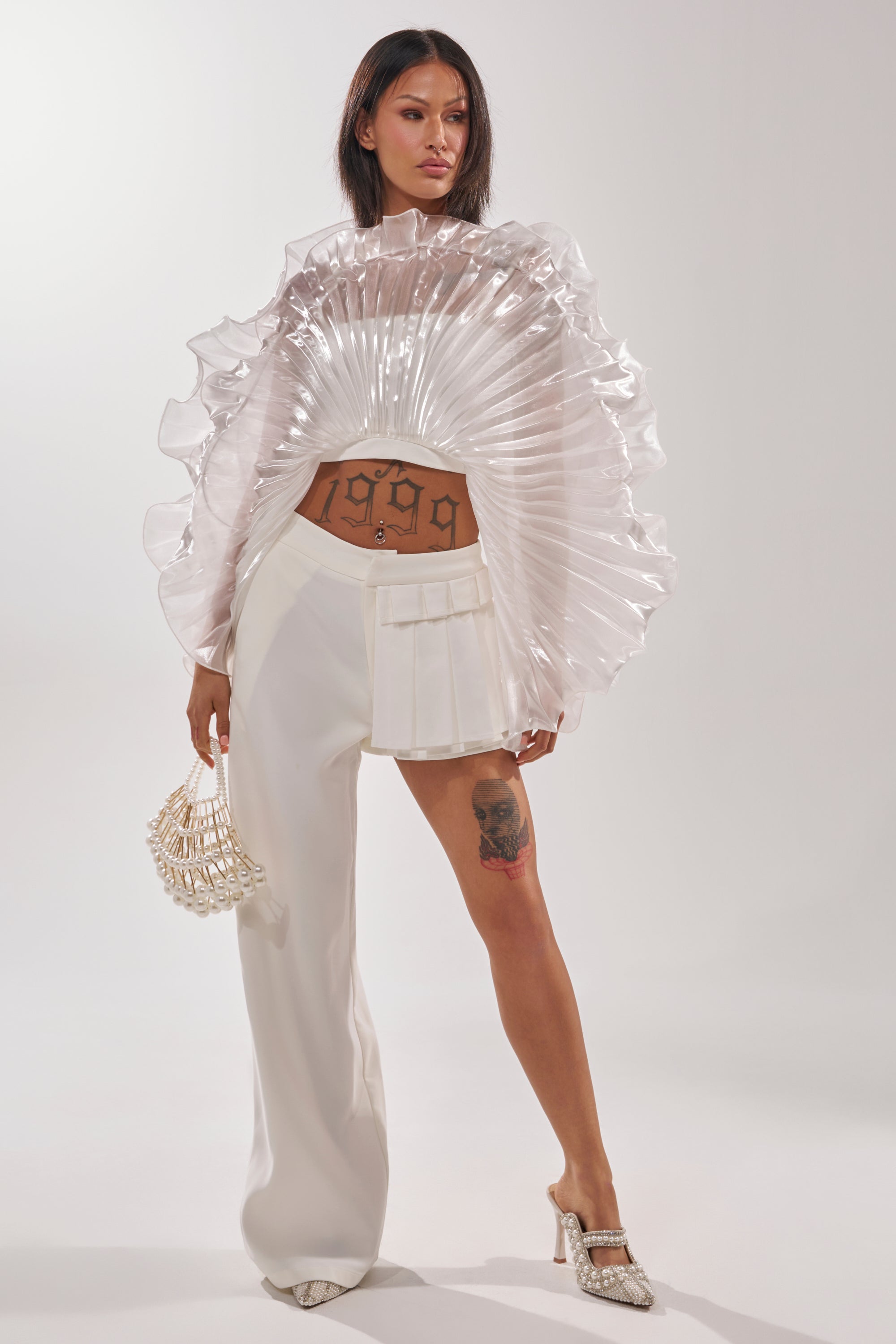 A woman stands confidently in fashion-forward attire, wearing the HOT AND COLD HYBRID PANT IN WHITE with an asymmetric silhouette—one pleated skirt leg and one wide-leg pant. She accessorizes with a pearl bag, heels, and showcases tattoos on her thigh and stomach. 0