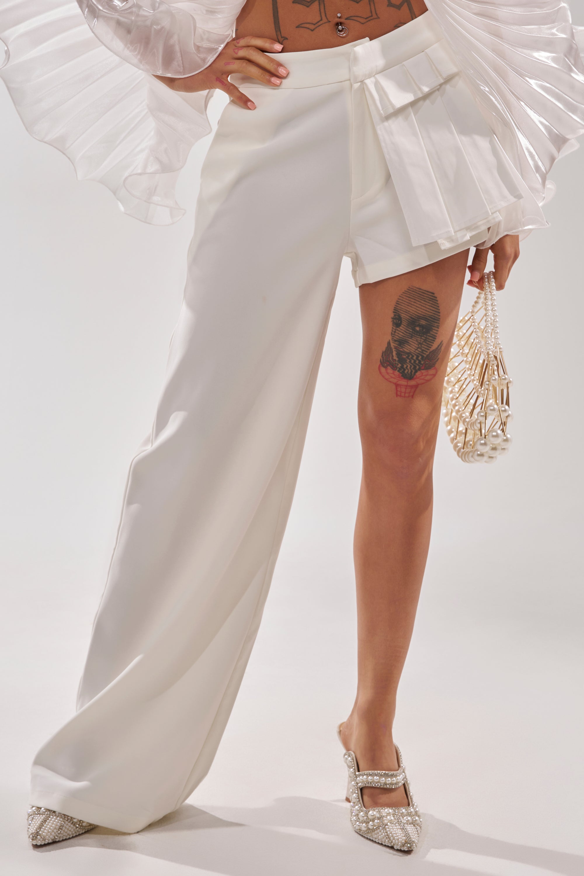 A person models the HOT AND COLD HYBRID PANT IN WHITE—one leg wide and asymmetric, the other a short. A skull tattoo is visible on their thigh, styled with a pearl handbag and sparkly heels. Only their lower half appears. 2