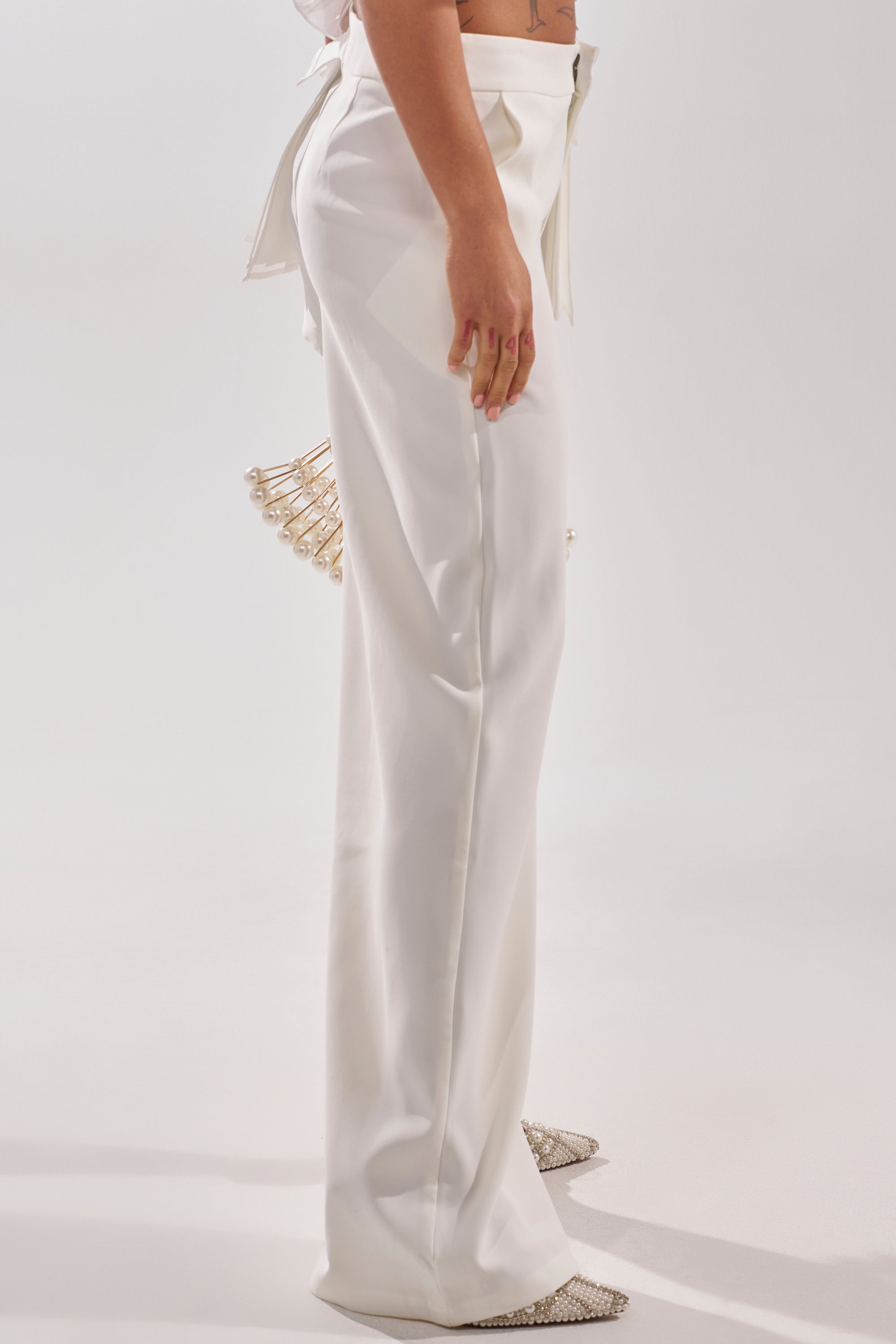 A person stands sideways against a white background, wearing HOT AND COLD HYBRID PANT IN WHITE with sparkly pointed-toe shoes. A decorative gold cage accessory peeks from the back of the pants, evoking modern luxury. 4