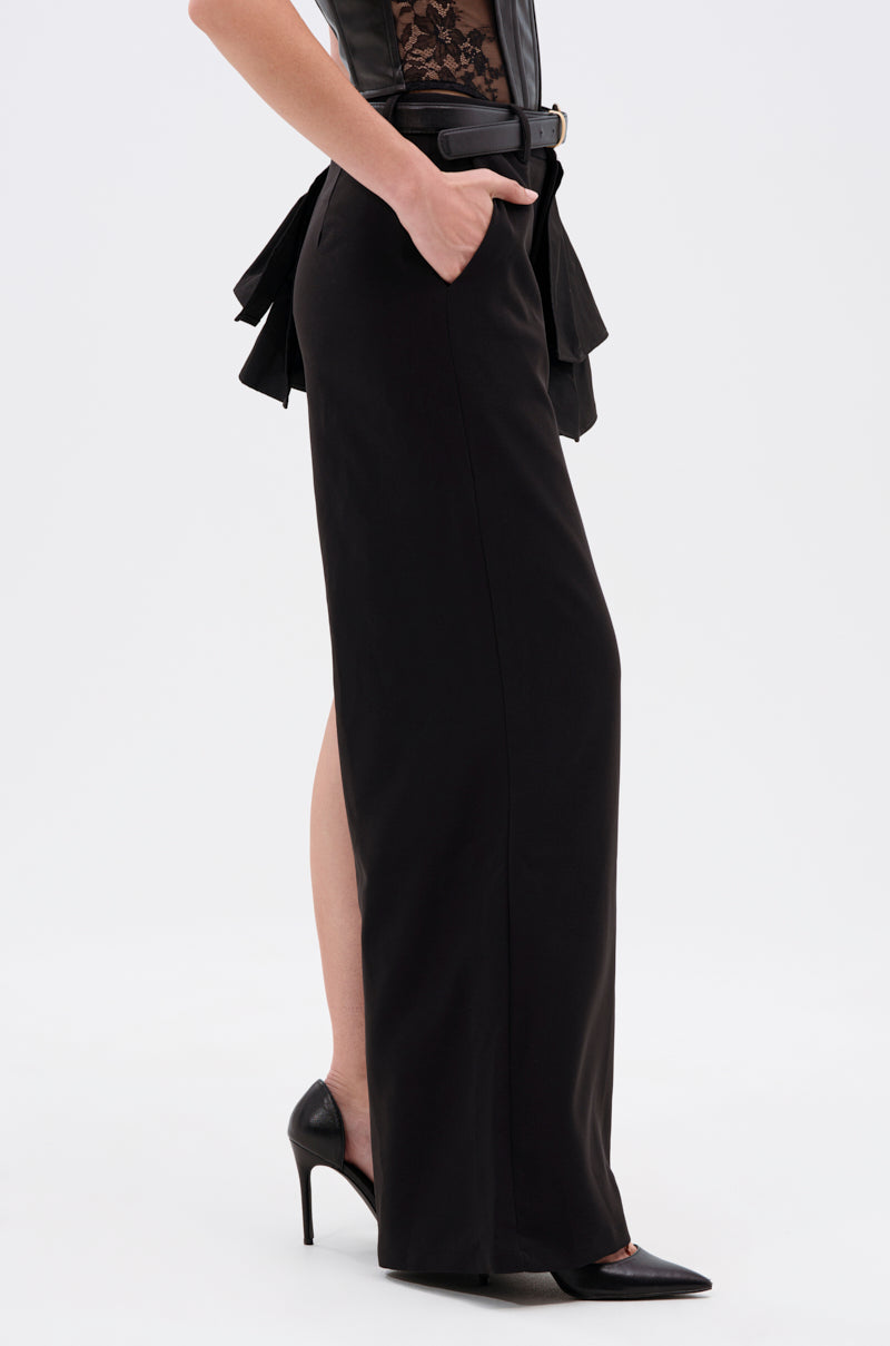 A person wears the HOT AND COLD HYBRID PANT with a side slit, paired with black high heels and a black lace top. A faux leather belt with a bow accents the waist, and their hand is in the pant pocket against a plain white background. 1