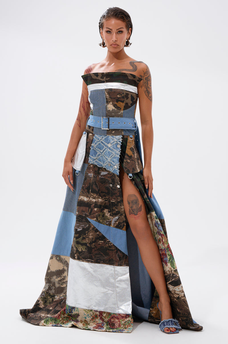 A woman in the STRAPLESS TRENCH MAXI DRESS WITH PATCHWORK DETAIL featuring blue, camouflage, and metallic silver fabrics, a wide blue belt, and blue open-toe heels poses confidently, revealing her thigh tattoo and multiple arm tattoos.
