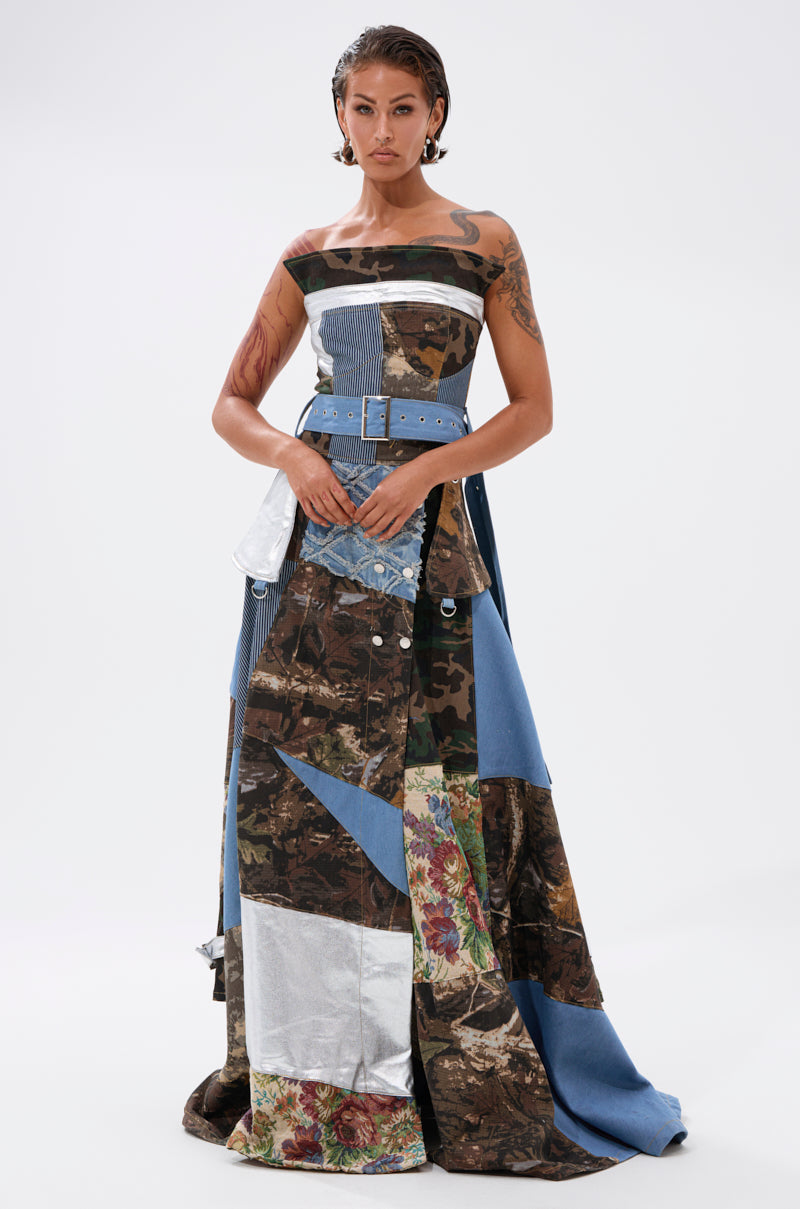 A woman stands confidently in the STRAPLESS TRENCH MAXI DRESS WITH PATCHWORK DETAIL—featuring denim, camouflage, floral, and solid panels—paired with a wide blue belt, set against a plain white background.