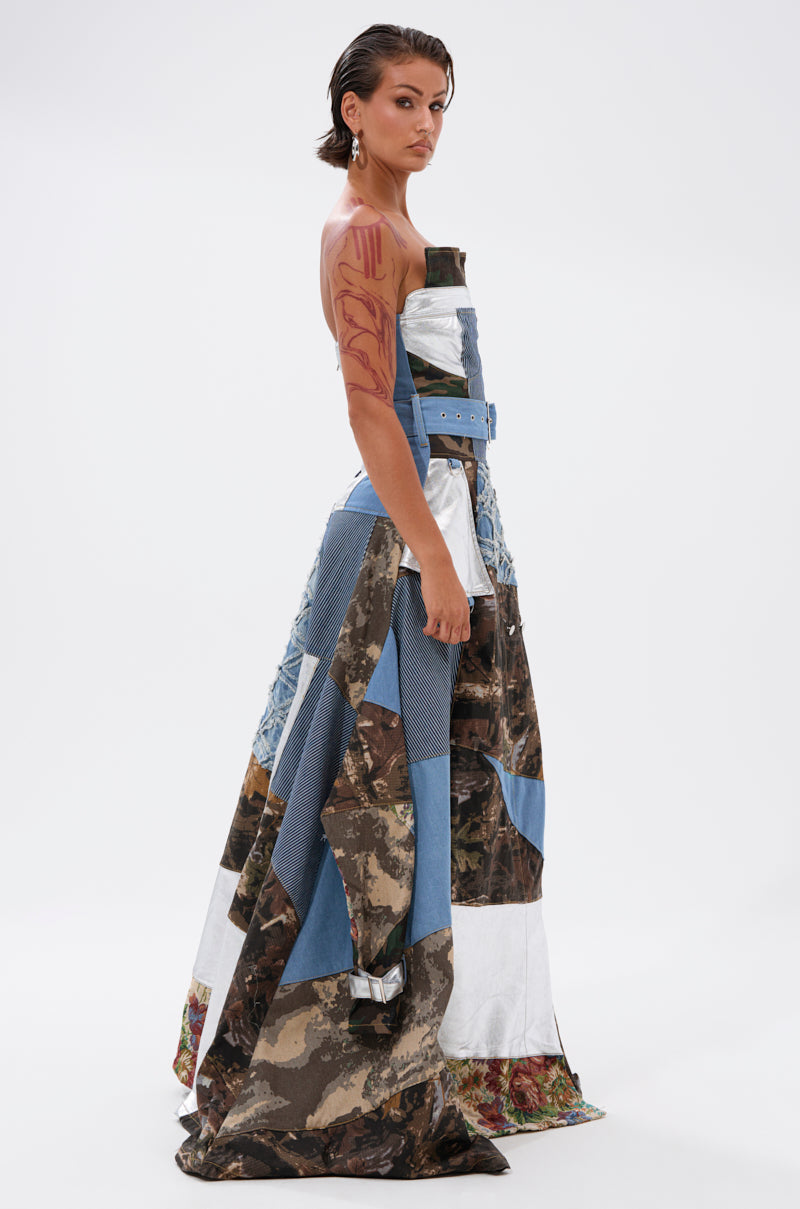 A person with slicked-back hair and body art poses sideways against a white background, wearing the STRAPLESS TRENCH MAXI DRESS WITH PATCHWORK DETAIL in denim, camouflage, and patterned fabrics.