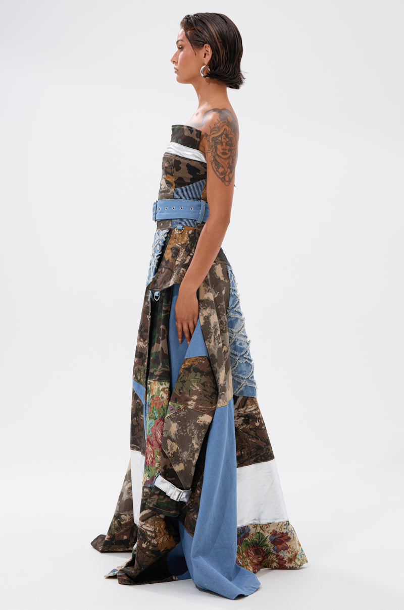 A woman with short dark hair and arm tattoos models the STRAPLESS TRENCH MAXI DRESS WITH PATCHWORK DETAIL—featuring camouflage, floral, and denim patterns with a blue belt—making a bold statement against a white background.