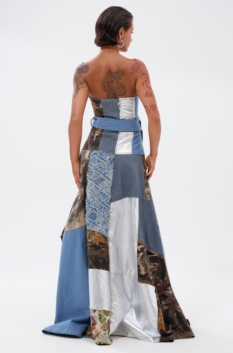 A woman with short hair and tattoos showcases the STRAPLESS TRENCH MAXI DRESS WITH PATCHWORK DETAIL, featuring denim, camouflage, silver metallic, and floral fabrics. She faces away, highlighting the back of this runway statement piece.