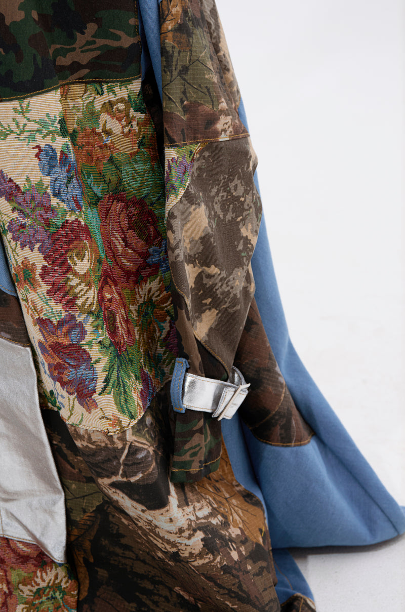 The STRAPLESS TRENCH MAXI DRESS WITH PATCHWORK DETAIL showcases floral tapestry, camouflage patterns, blue fabric, and a silver buckle in a standout design, perfect for making a runway statement against a plain white background.