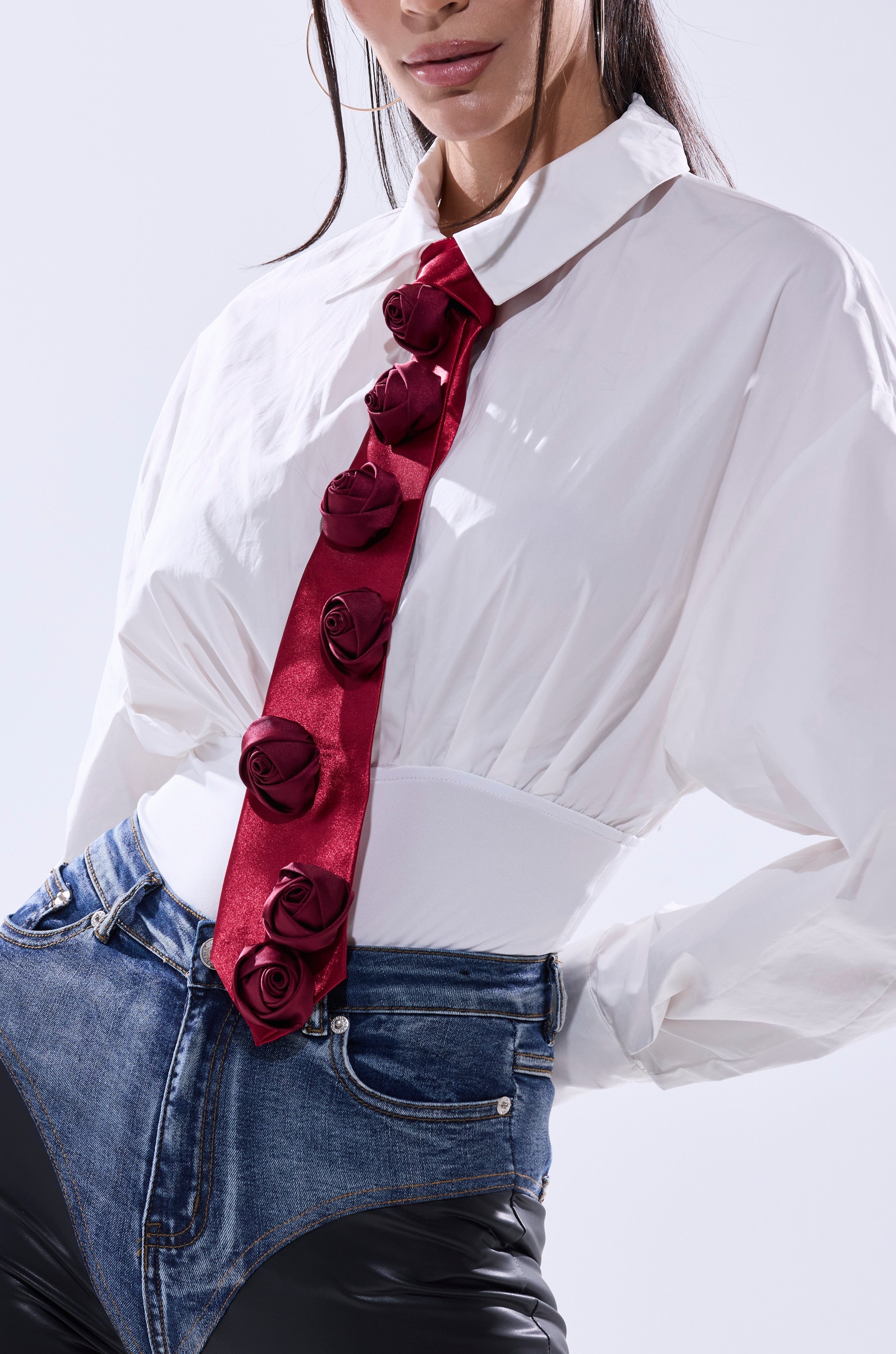 Wearing the ROSEY TIE—a red tie with fabric roses—a woman pairs it with a white shirt and high-waisted denim pants featuring black leather sections. Her hair partially covers her face.