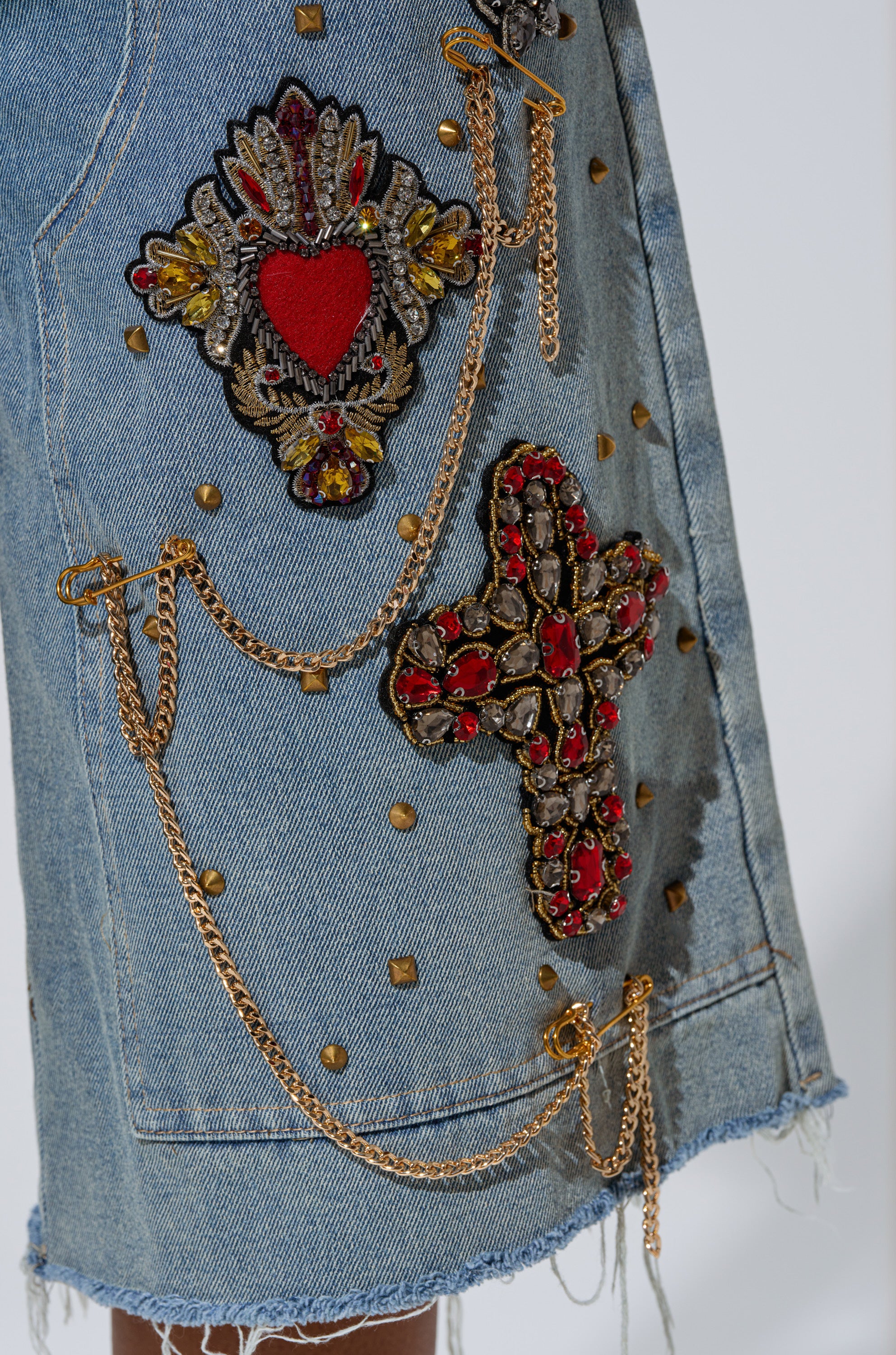 Close-up of the COOL GIRL LIKE ME BERMUDA SHORT in light-wash denim, featuring gold-tone studs, chains, ornate red and gold embroidered heart and cross patches, and a stylish frayed hem. 9