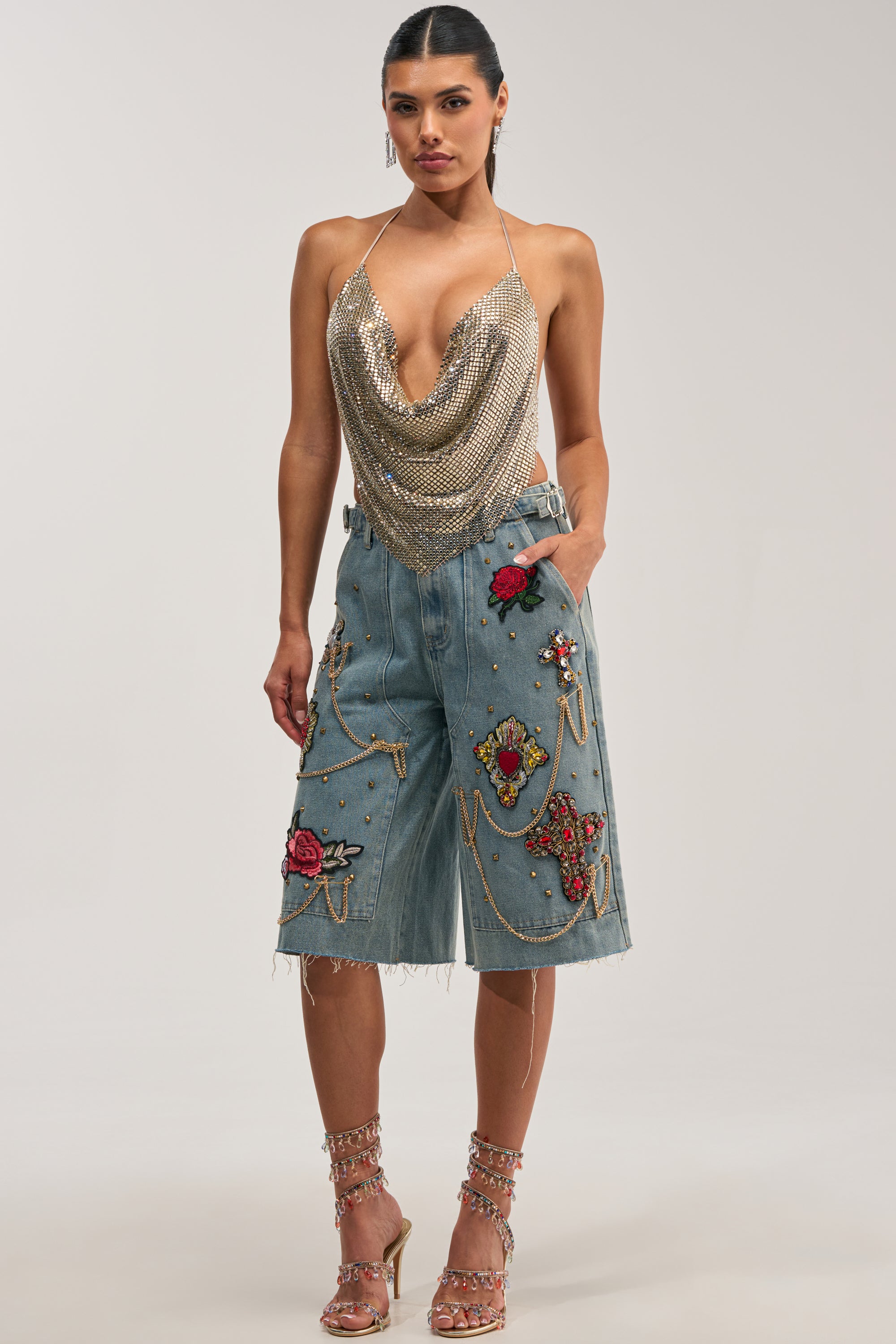 A woman models the COOL GIRL LIKE ME BERMUDA SHORT, an embellished denim Bermuda short adorned with rose and religious-themed patches, gold-tone studs, and gold chains. She pairs it with a gold chainmail halter top and heeled embellished sandals, capturing effortless street style against a plain light gray background.