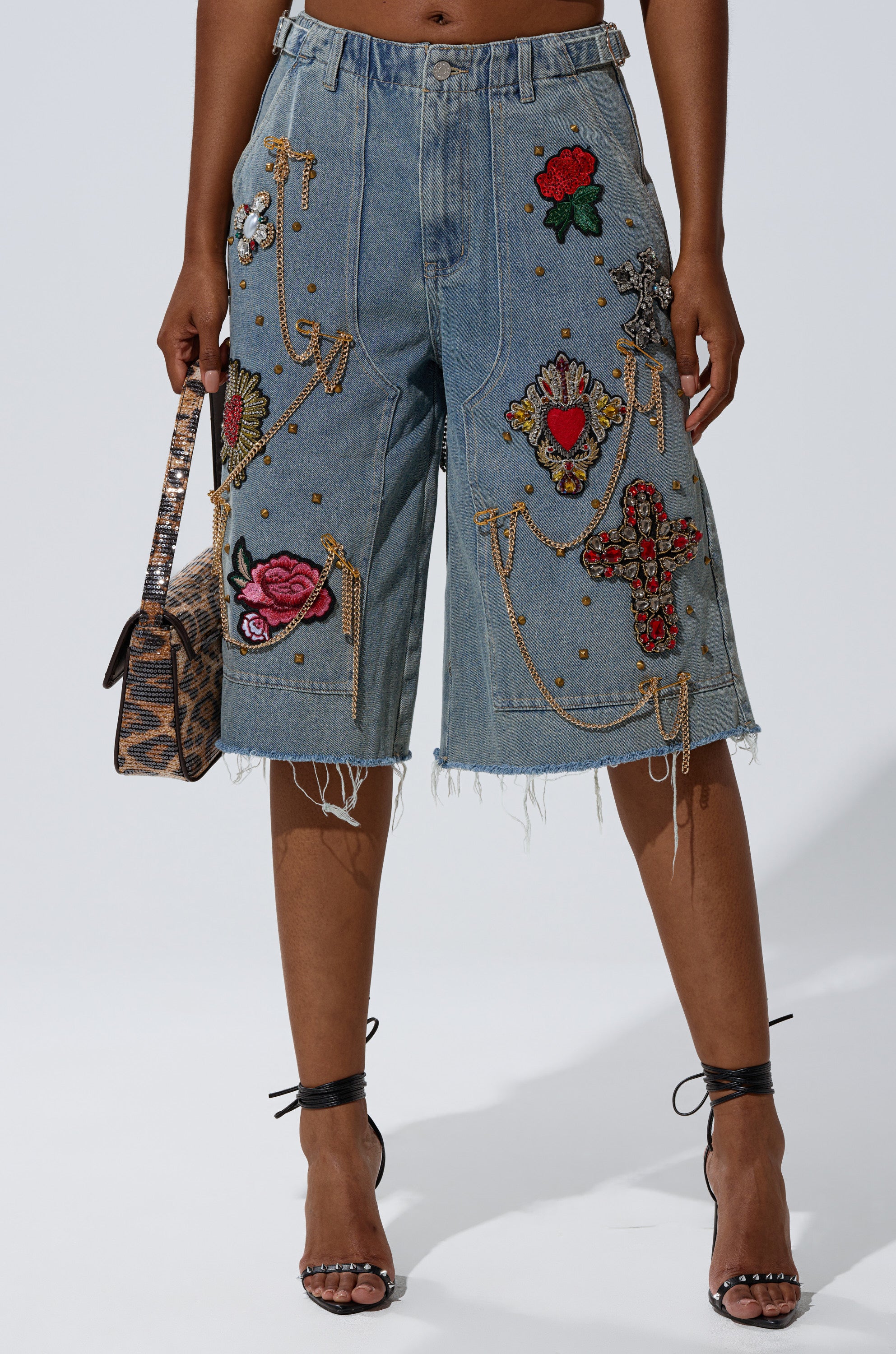 A person wearing the COOL GIRL LIKE ME BERMUDA SHORT—knee-length, light-wash denim with colorful patches, beads, gold studs, and chains—paired with a patterned handbag and black strappy heels. 1