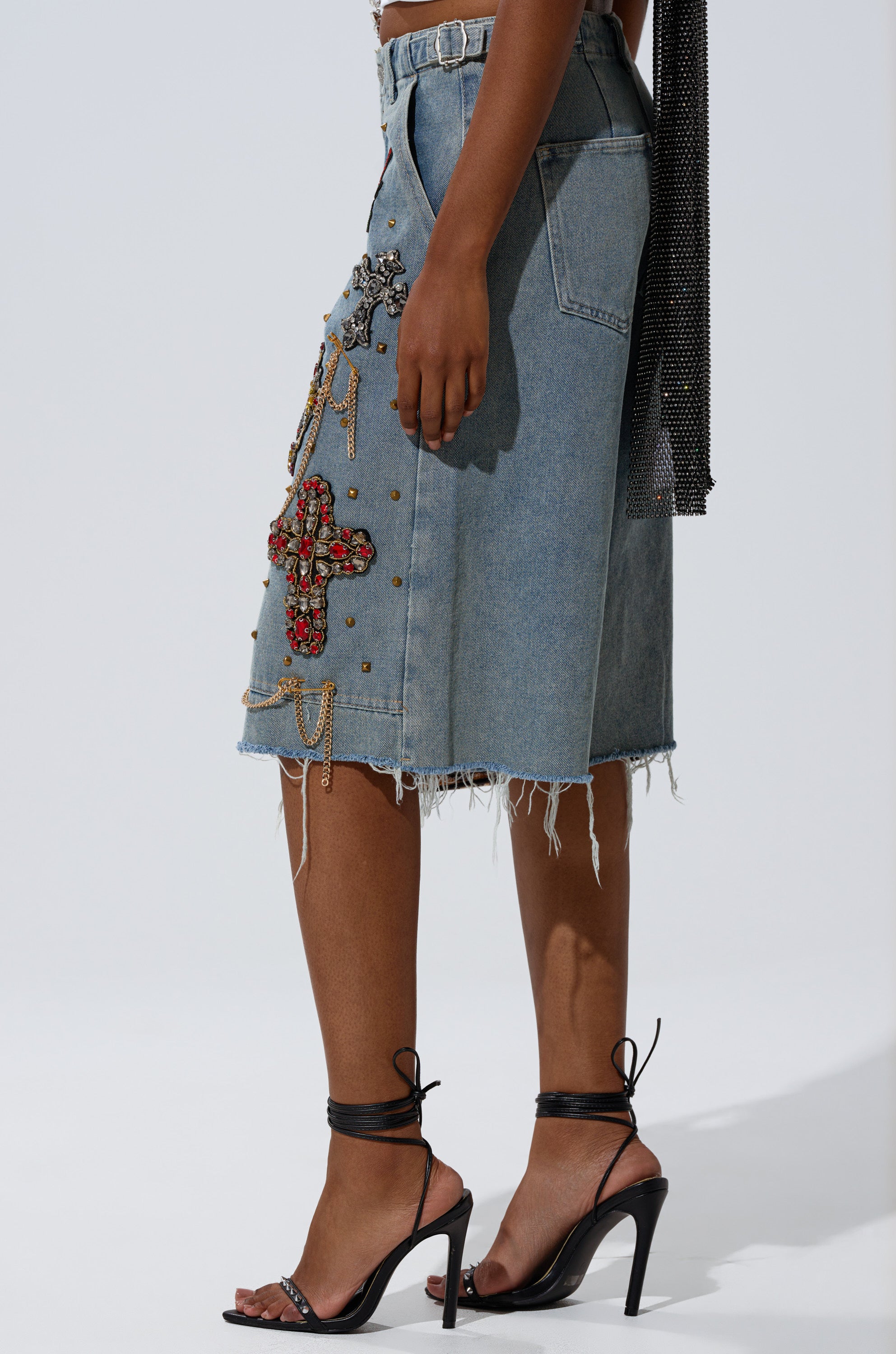 A person wears the COOL GIRL LIKE ME BERMUDA SHORT—knee-length, light-wash denim with a frayed hem, beaded crosses, and gold-tone studs—paired with black high-heeled sandals against a plain, well-lit background. 2