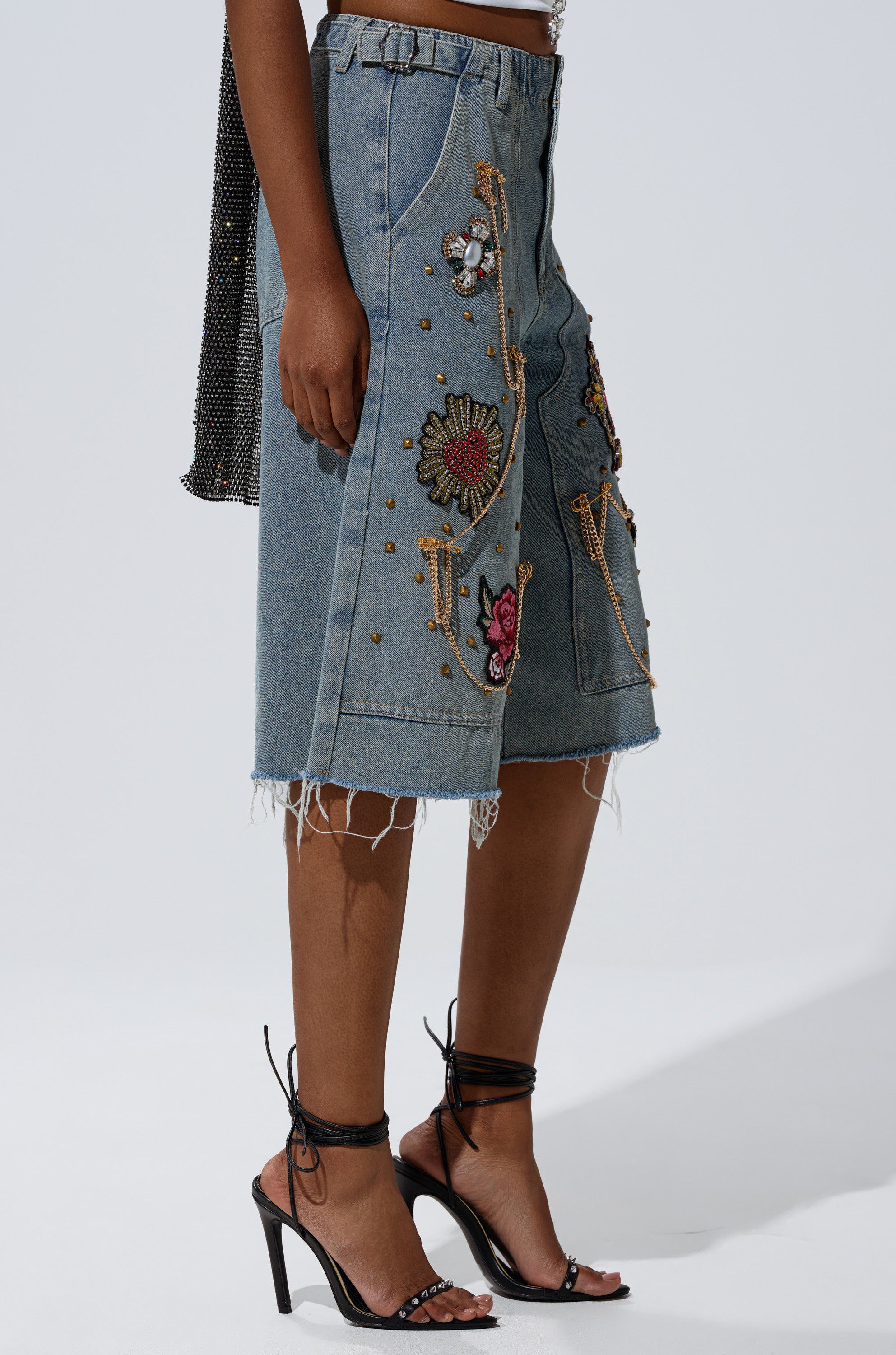 Someone wears the COOL GIRL LIKE ME BERMUDA SHORT—light-wash denim with vibrant embroidered and beaded patches—paired with black high-heeled sandals, standing against a plain background. 3