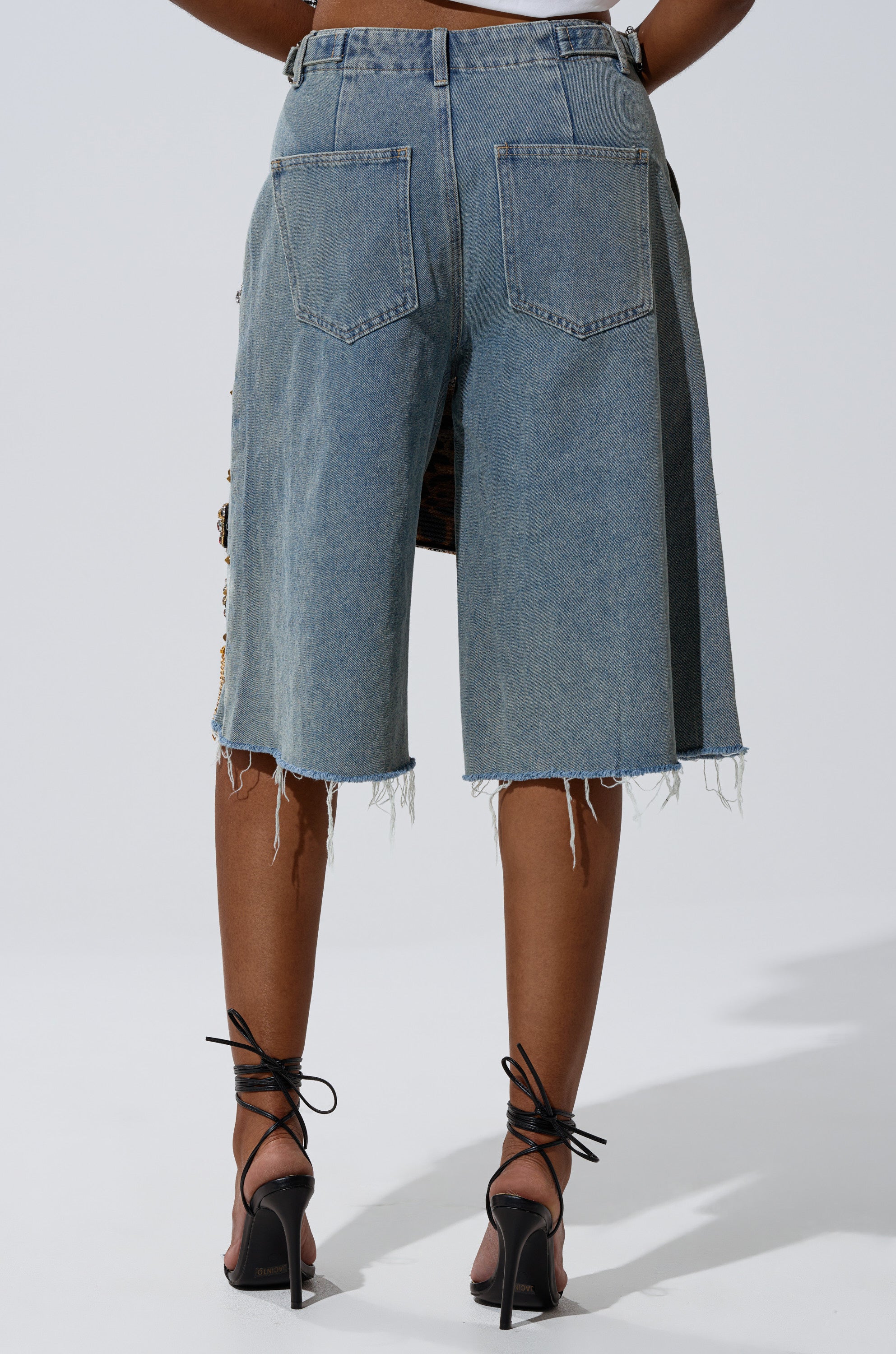 A person faces away, wearing the COOL GIRL LIKE ME BERMUDA SHORT—light-wash denim with a mid-rise fit, raw uneven hems, and two back pockets—paired with black strappy high-heeled sandals. 4