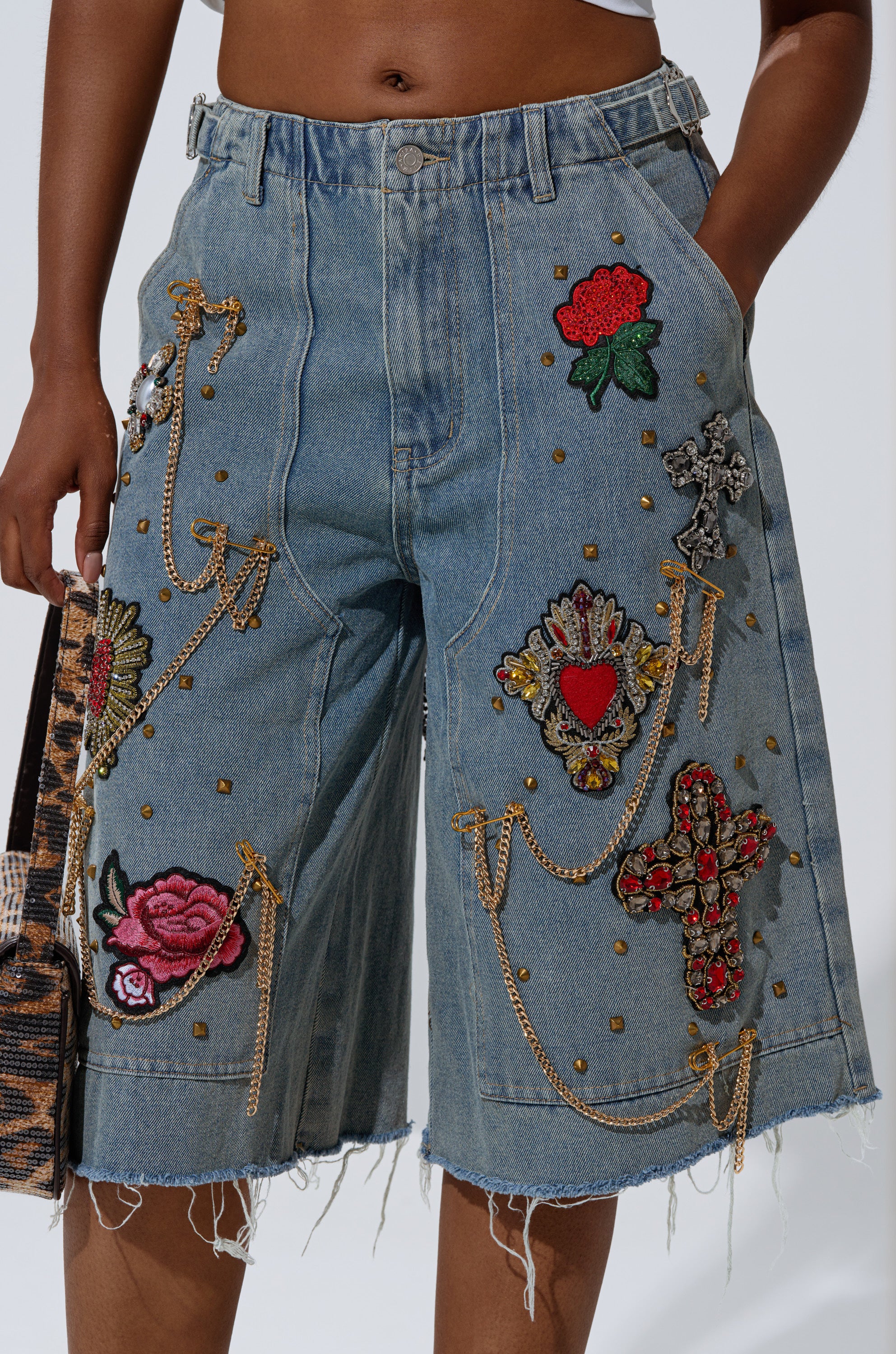A person wearing COOL GIRL LIKE ME BERMUDA SHORT, knee-length distressed denim with frayed hems, colorful embroidered patches, gold-tone studs and chains, featuring bold rose, heart, and cross designs. 8