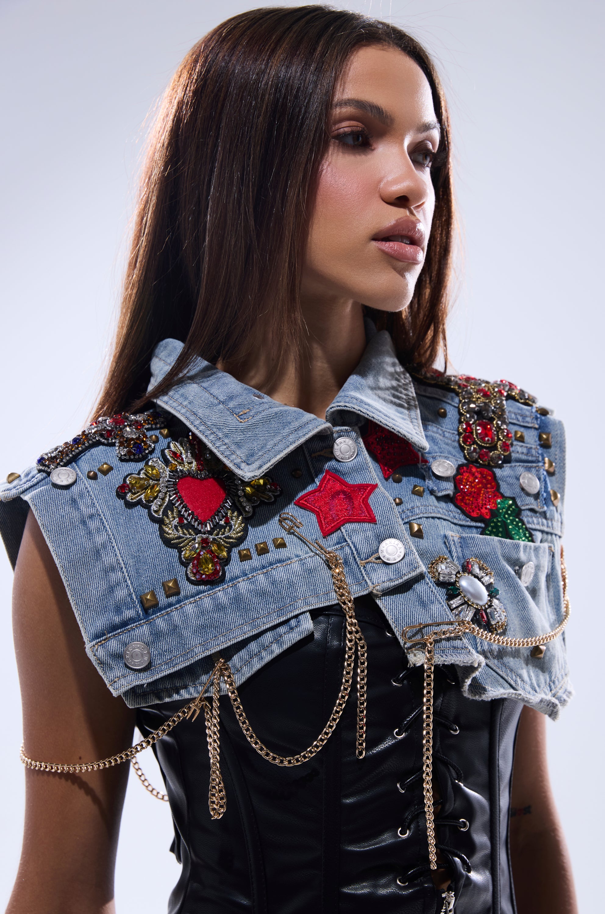 A woman with straight brown hair wears the STYLE ME UP DENIM COLLAR—a black corset top and a studded vest with colorful patches, beads, and gold chains—looking to the side with a neutral expression against a light background.