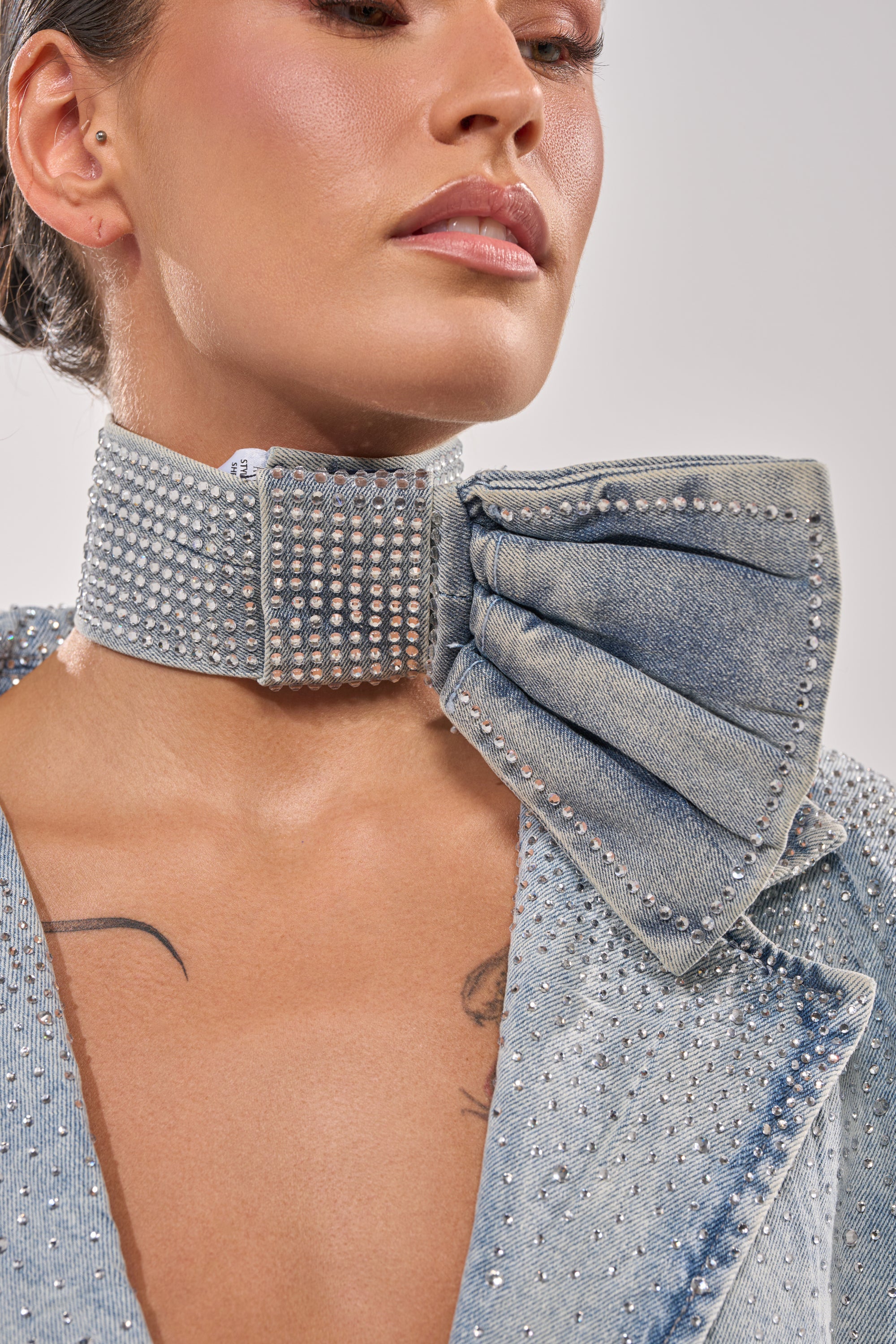 A woman wears the WITH A BOW DENIM NECKLACE, a denim choker with rhinestones and an oversized bow, paired with a matching embellished jacket. Her makeup is subtle and part of a chest tattoo is visible. 0