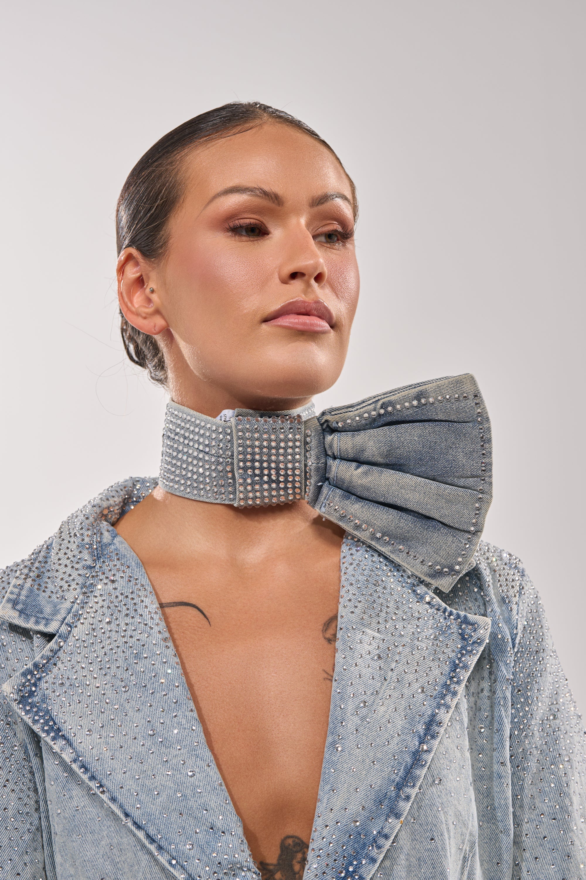 A woman wears a studded light denim jacket with the WITH A BOW DENIM NECKLACE—a matching denim bow choker adorned with rhinestones—confidently looking to the side against a plain background, making the necklace the focal point. 1