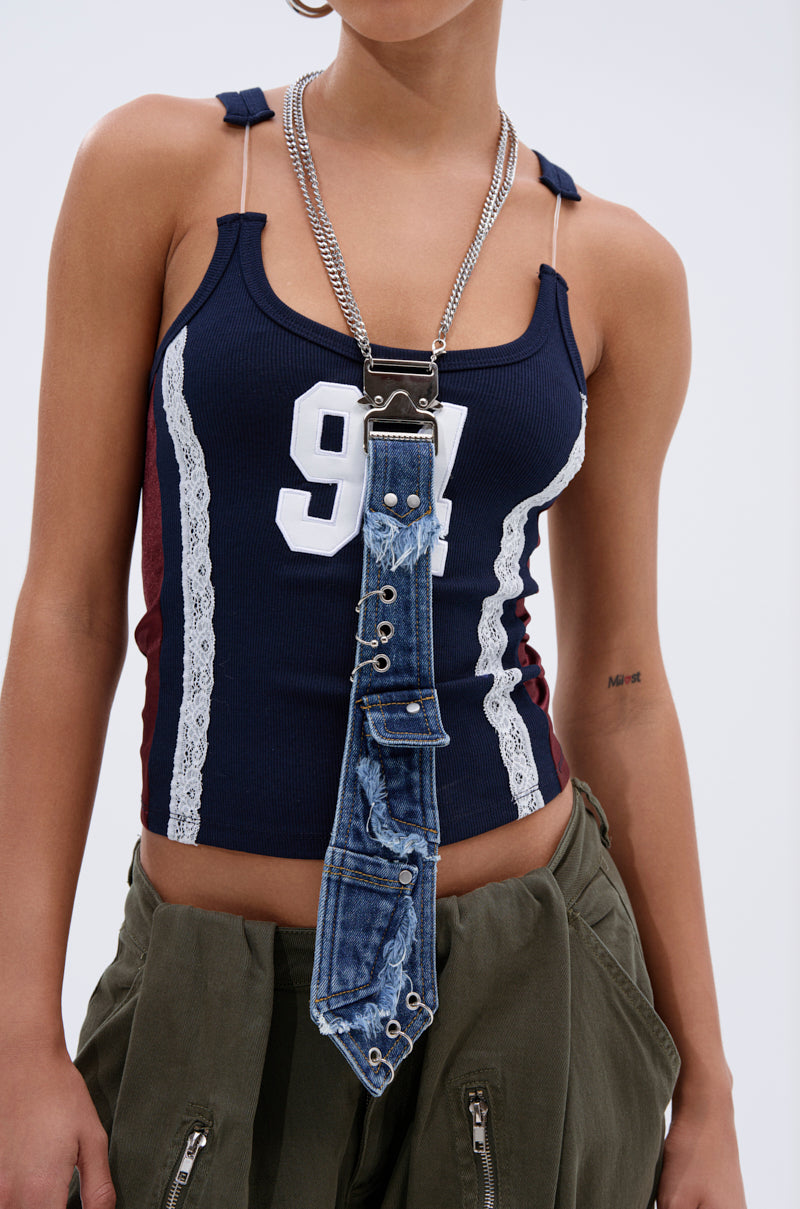 A person wears a navy tank top with "94" and lace trim, olive green pants, and the MIXED FEELINGS DENIM TIE adorned with metal grommets, hanging from a silver chain necklace. A small tattoo is visible on their upper left arm.