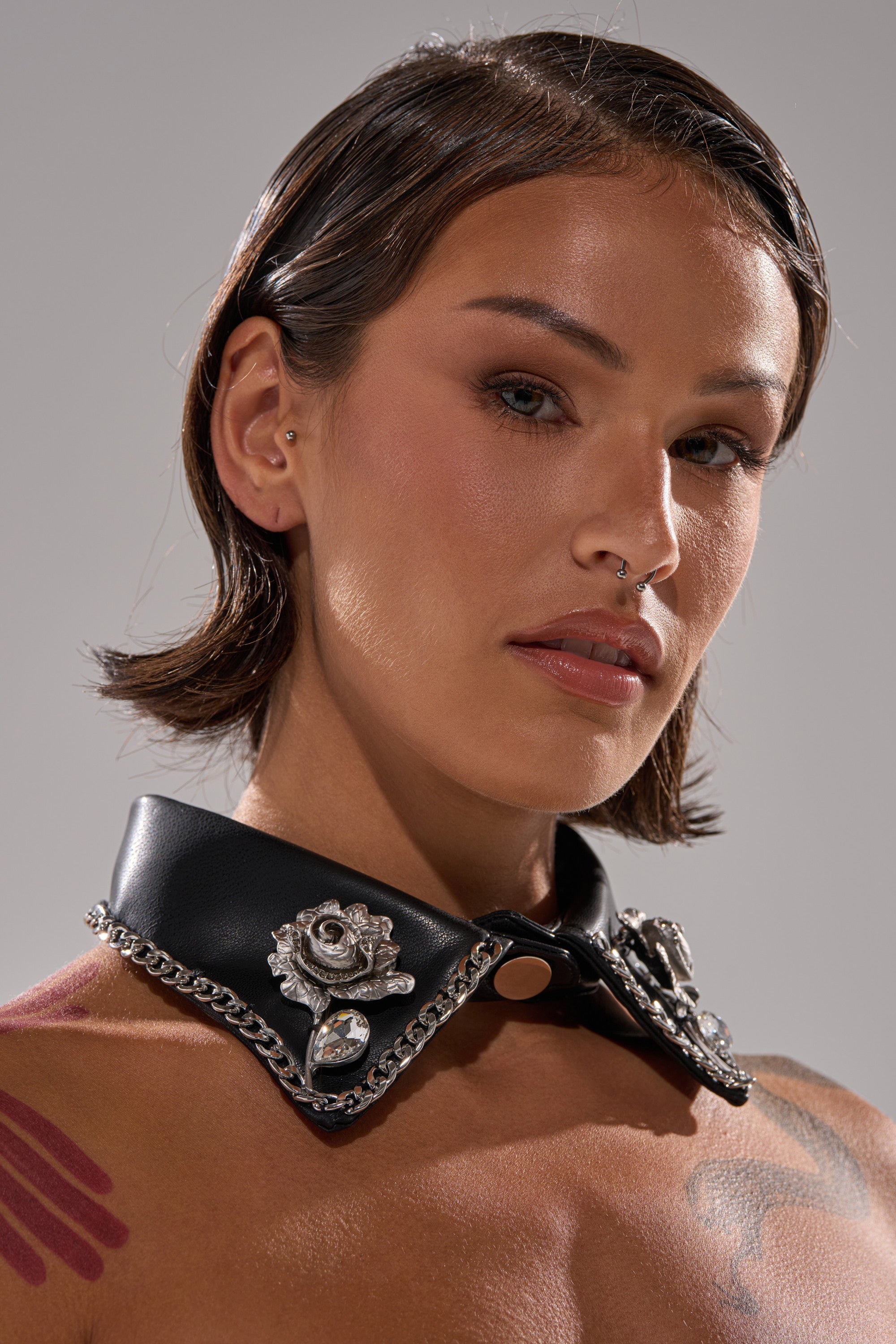 A person with short brown hair, light makeup, and a septum piercing wears the GRACEY PU COLLAR NECKLACE featuring metal roses and chains. The plain background is well-lit. 0