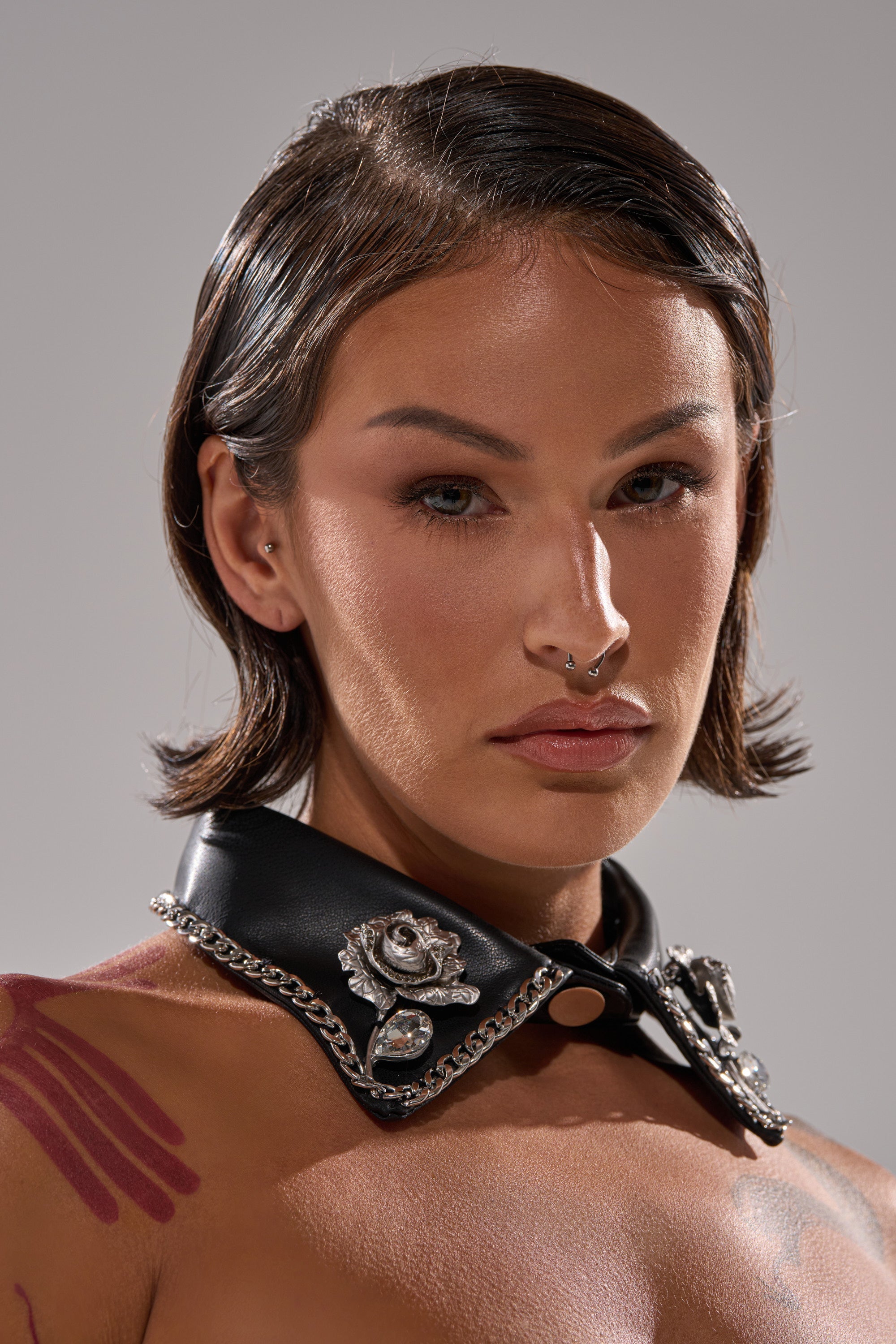 A person with short, dark hair and a septum piercing wears the GRACEY PU COLLAR NECKLACE, a black faux leather accessory with metal and jeweled details. They have a neutral expression and abstract pink markings on their shoulders. 2
