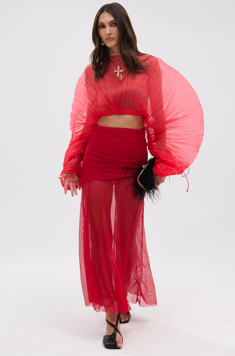 A woman in a sheer, bright red top with dramatic pleated sleeves and the EASY ENOUGH RED MESH MIDI SKIRT holds a black feathered clutch while standing against a plain white background. 1