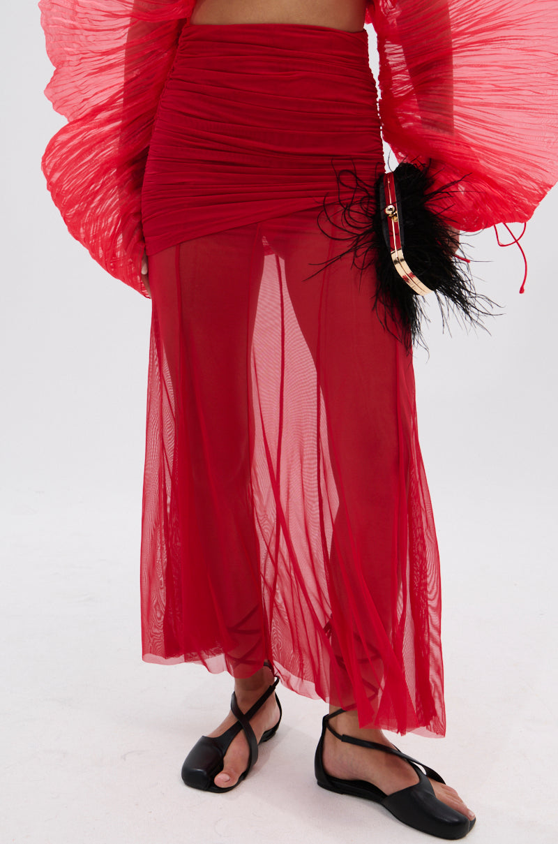 A person wears the EASY ENOUGH RED MESH MIDI SKIRT with a matching sheer red top with pleated sleeves, black flat sandals, and holds a small black feathered bag with gold detailing. The background is white. 3