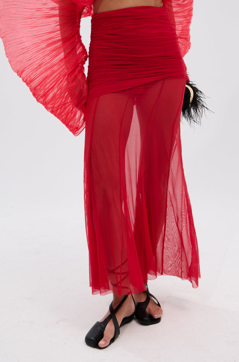 Person wears the EASY ENOUGH RED MESH MIDI SKIRT with a matching pleated, wide-sleeve top, black sandals, and holds a small black feathered accessory against a plain white background. 4