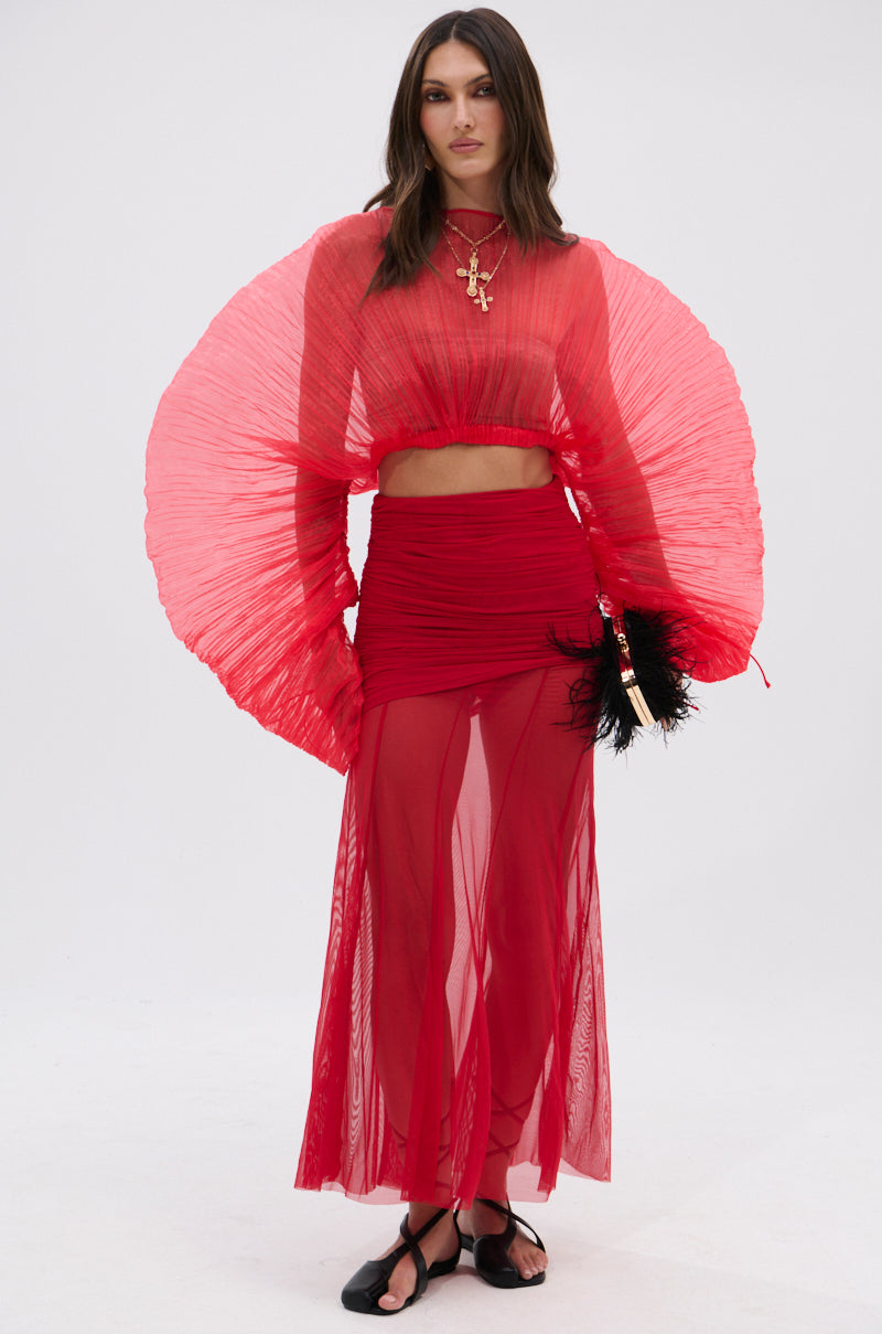 A woman poses against a plain background wearing the EASY ENOUGH RED MESH MIDI SKIRT as part of a two-piece set with dramatic pleated sleeves, paired with layered gold necklaces, black sandals, and a black feathered clutch. 7