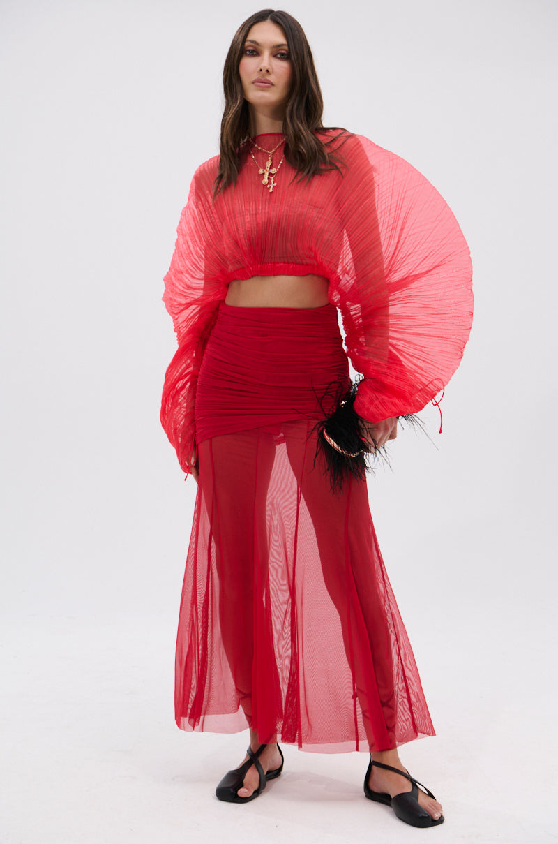 A woman poses against a plain white background in the EASY ENOUGH RED MESH MIDI SKIRT, styled with large pleated sleeves, black sandals, a feathered black clutch, and layered necklaces. 8