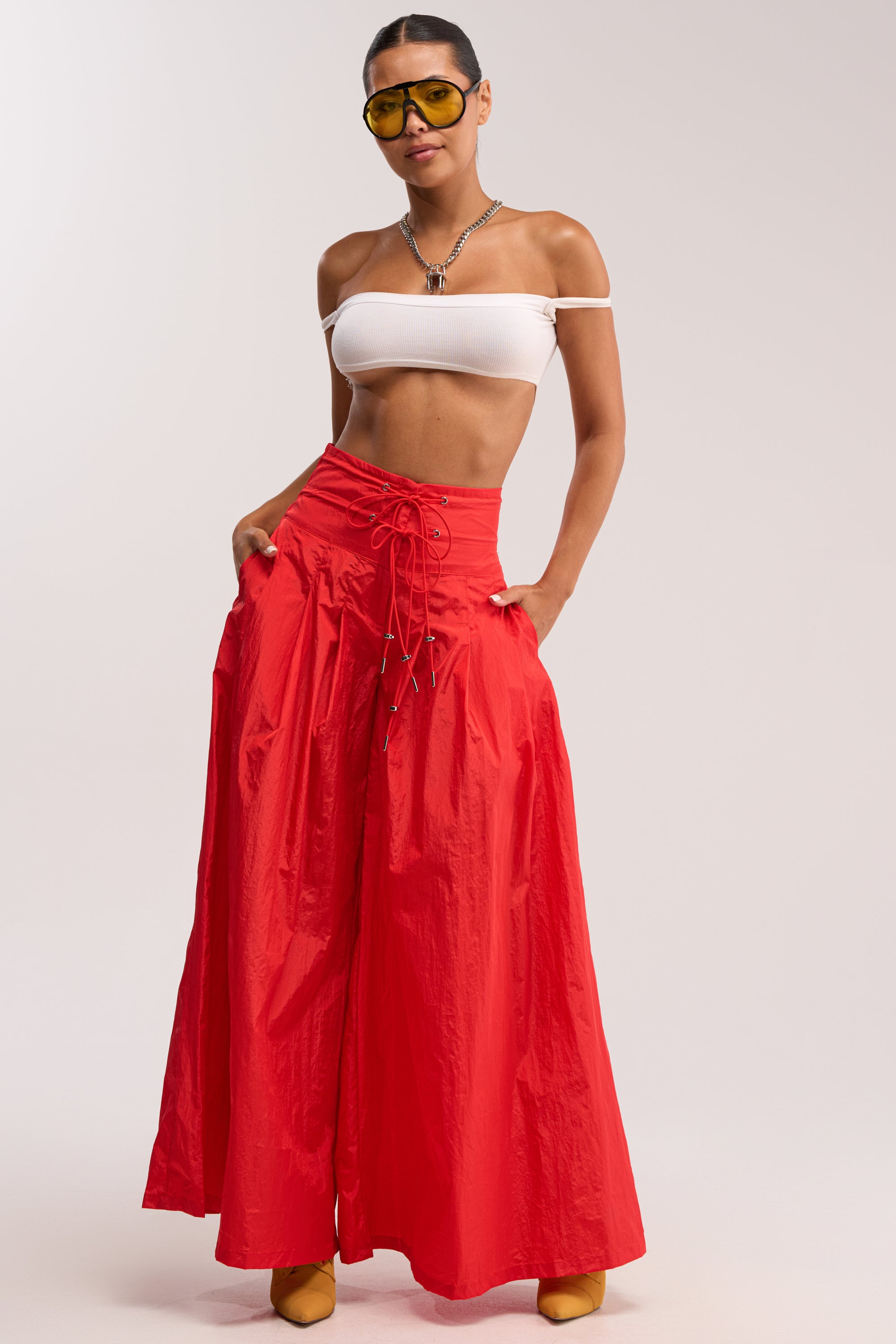 A person wearing a white off-shoulder crop top, DANCE THE NIGHT AWAY PALAZZO PANT IN RED, yellow sunglasses, yellow shoes, and a silver necklace stands with hands in pockets against a plain background.