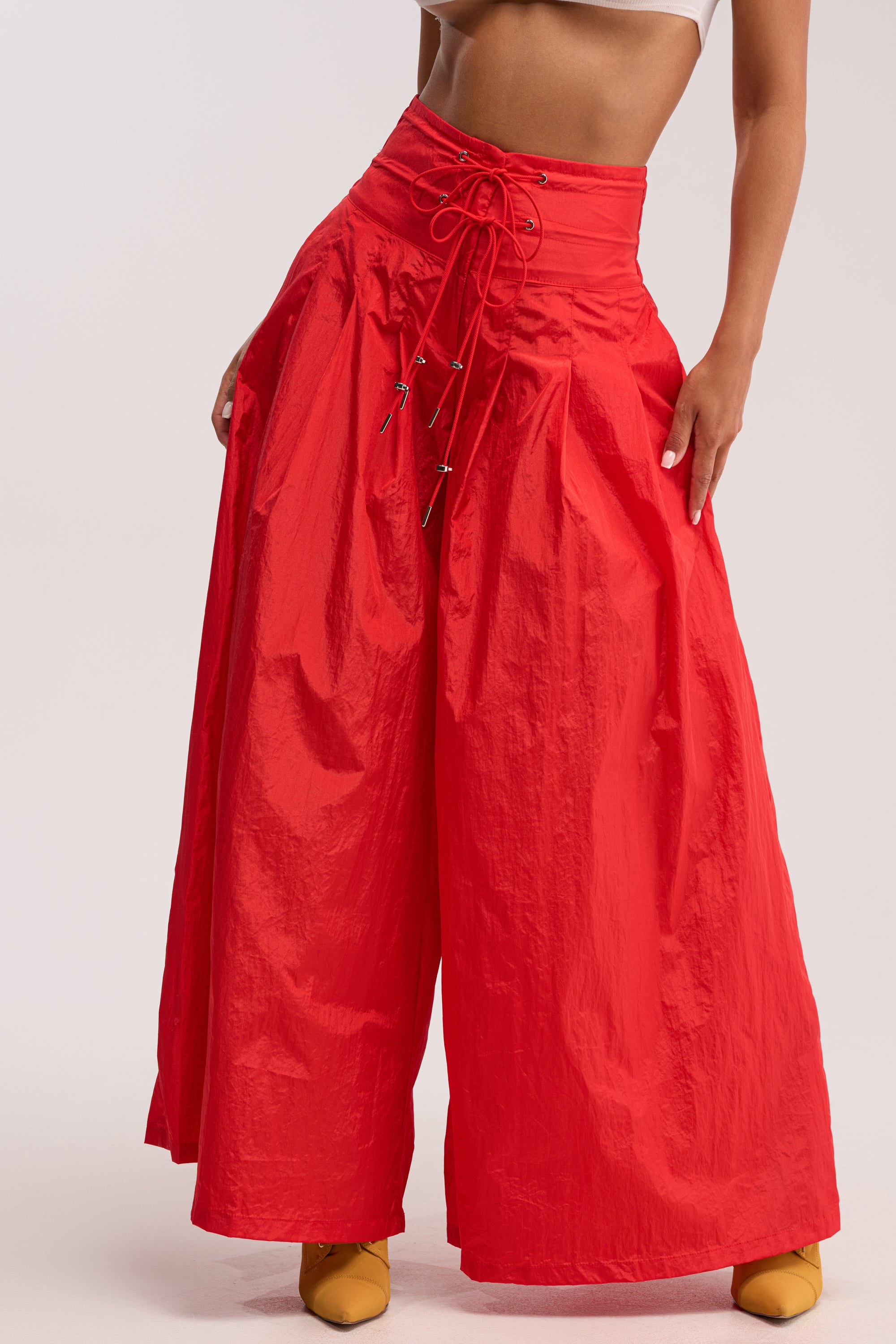 Wearing the DANCE THE NIGHT AWAY PALAZZO PANT IN RED—ultra-high waist, wide-leg, lace-up utility detail—a person pairs them with a white crop top and yellow pointed-toe shoes, standing against a plain background.