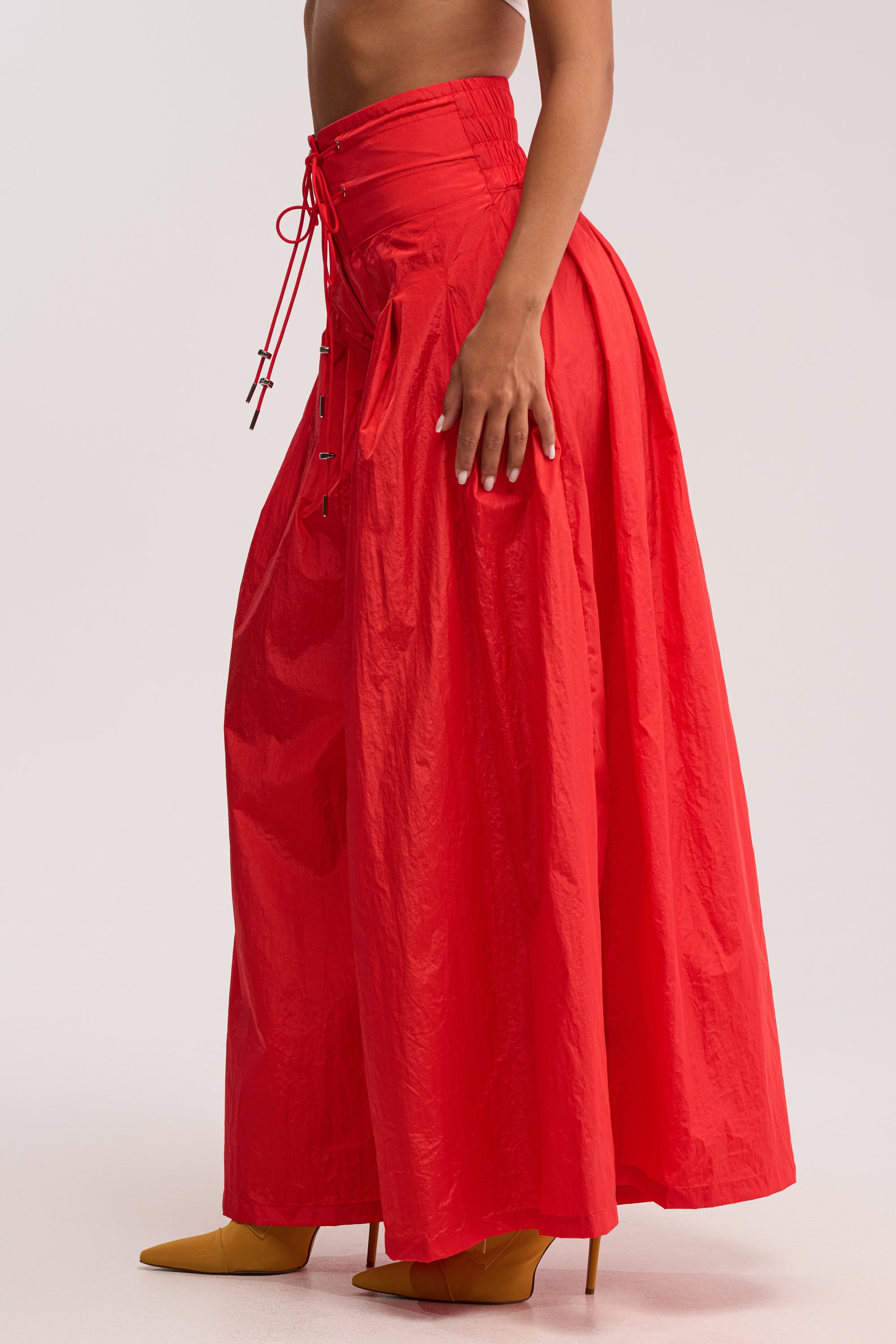 A person wearing the DANCE THE NIGHT AWAY PALAZZO PANT IN RED—bright, wide-leg style with a fitted lace-up waistband—paired with mustard yellow pointed-toe high heels.
