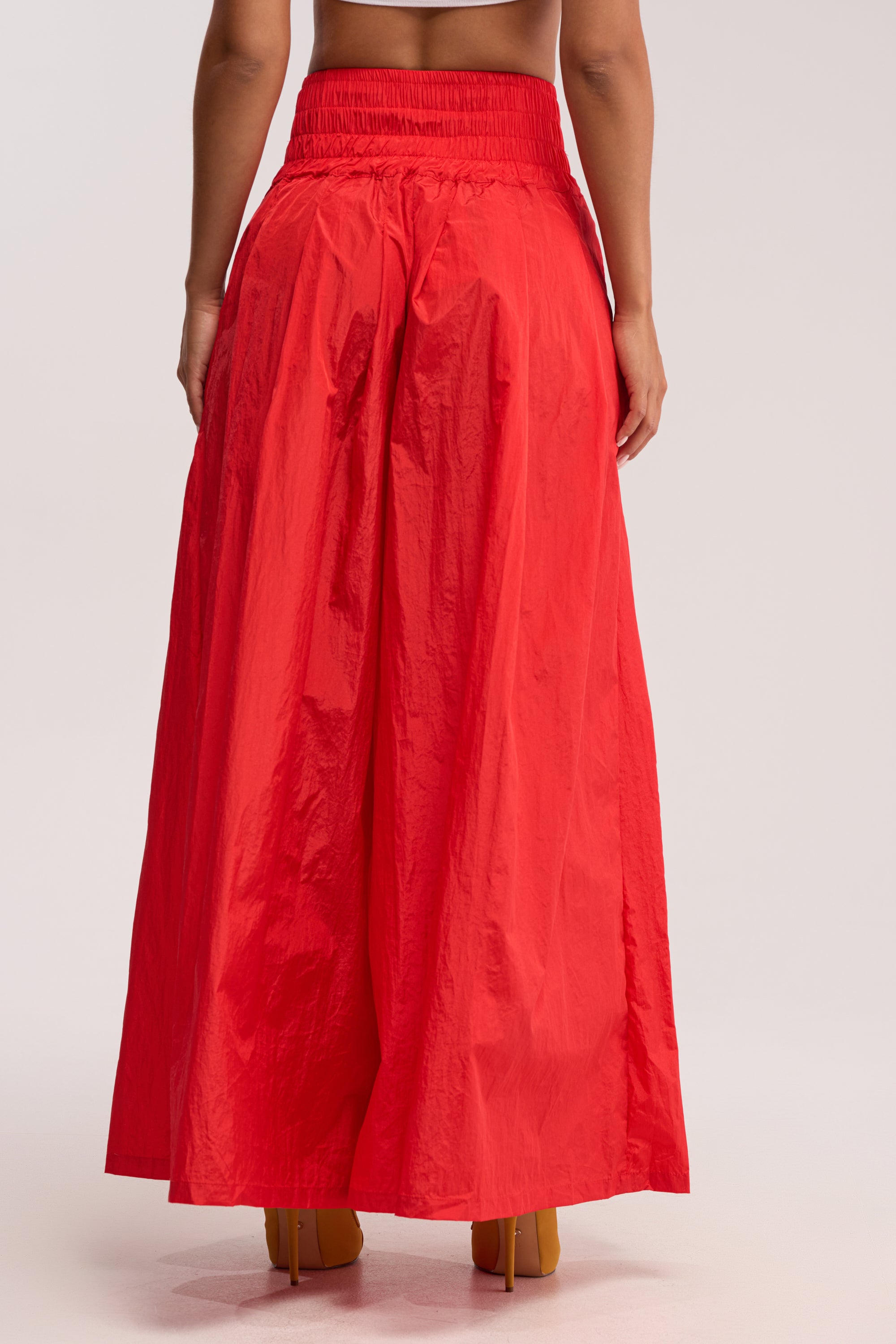 A person wearing the DANCE THE NIGHT AWAY PALAZZO PANT IN RED, featuring a high waist, wide-leg fit, and adjustable lace-up waistband, stands facing away from the camera against a light background. Only the lower back and legs are visible.