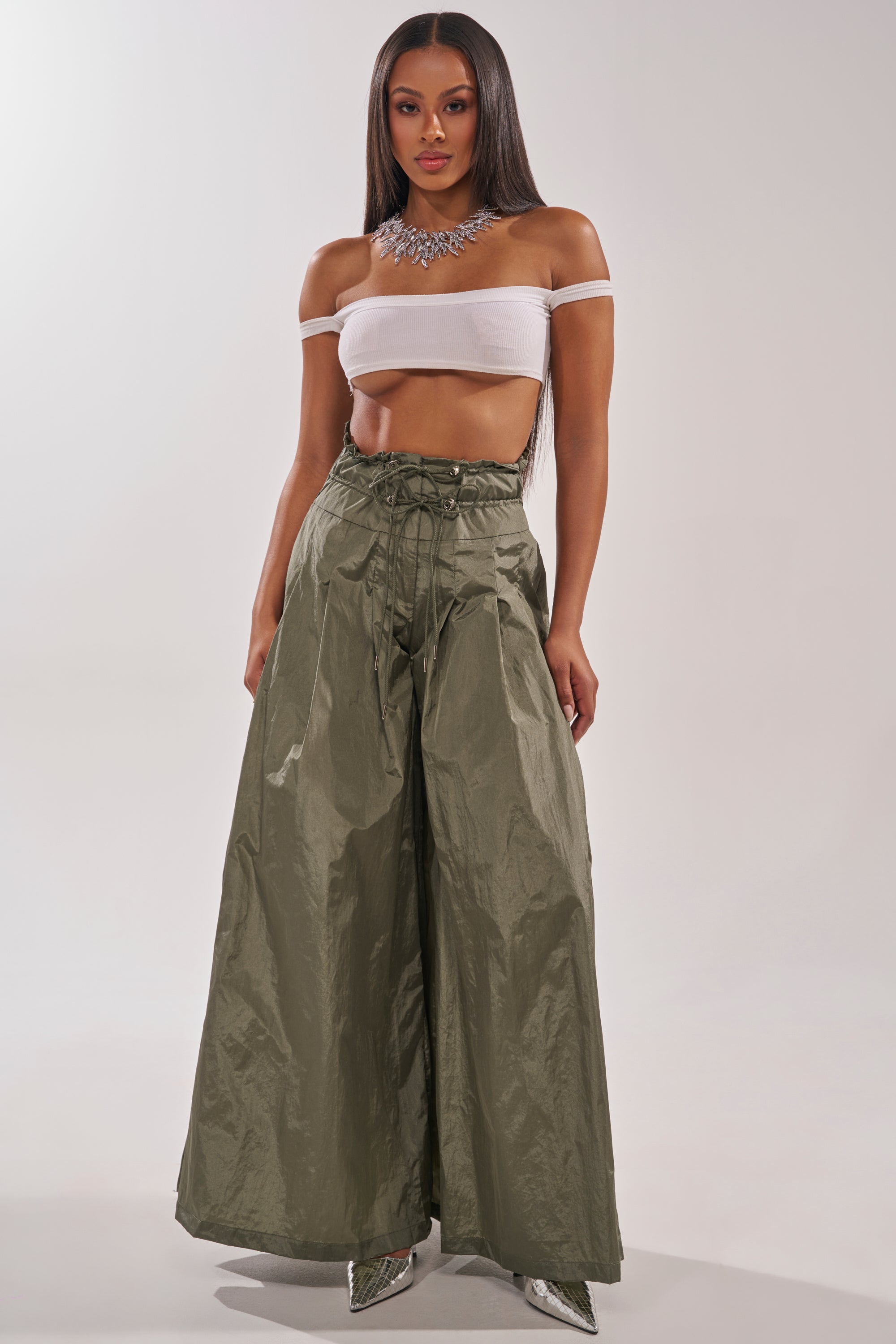 A woman stands confidently in the DANCE THE NIGHT AWAY PALAZZO PANT—high-rise olive green wide-leg utility pants with a lace-up front—paired with a white off-the-shoulder crop top, silver pointed-toe shoes, and a statement necklace against a plain background. 0