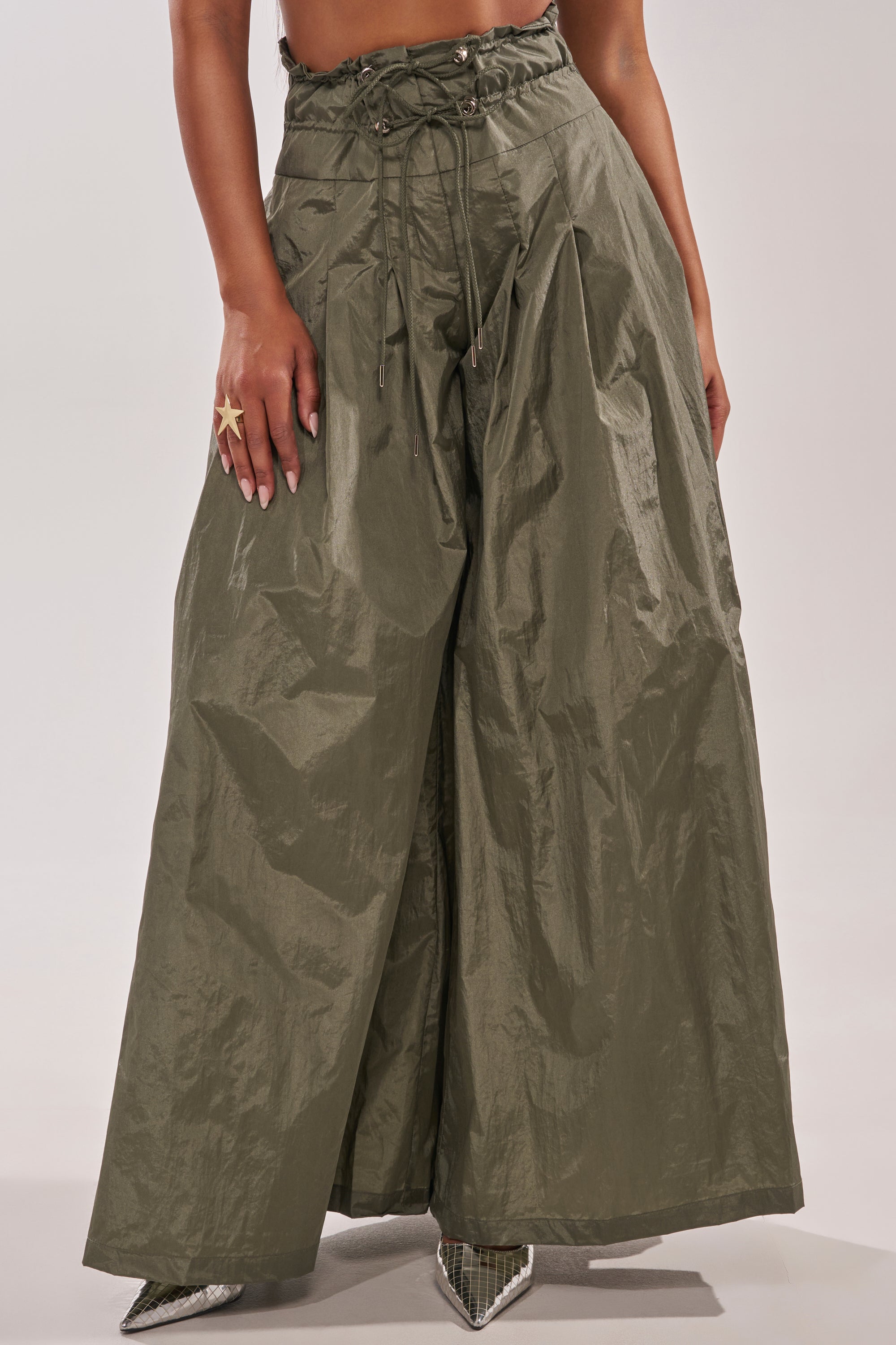 A person wears the DANCE THE NIGHT AWAY PALAZZO PANT—olive green, high-rise, wide-leg pants with a drawstring waist and pleats—paired with pointed-toe heels. Only the lower body is visible. 1