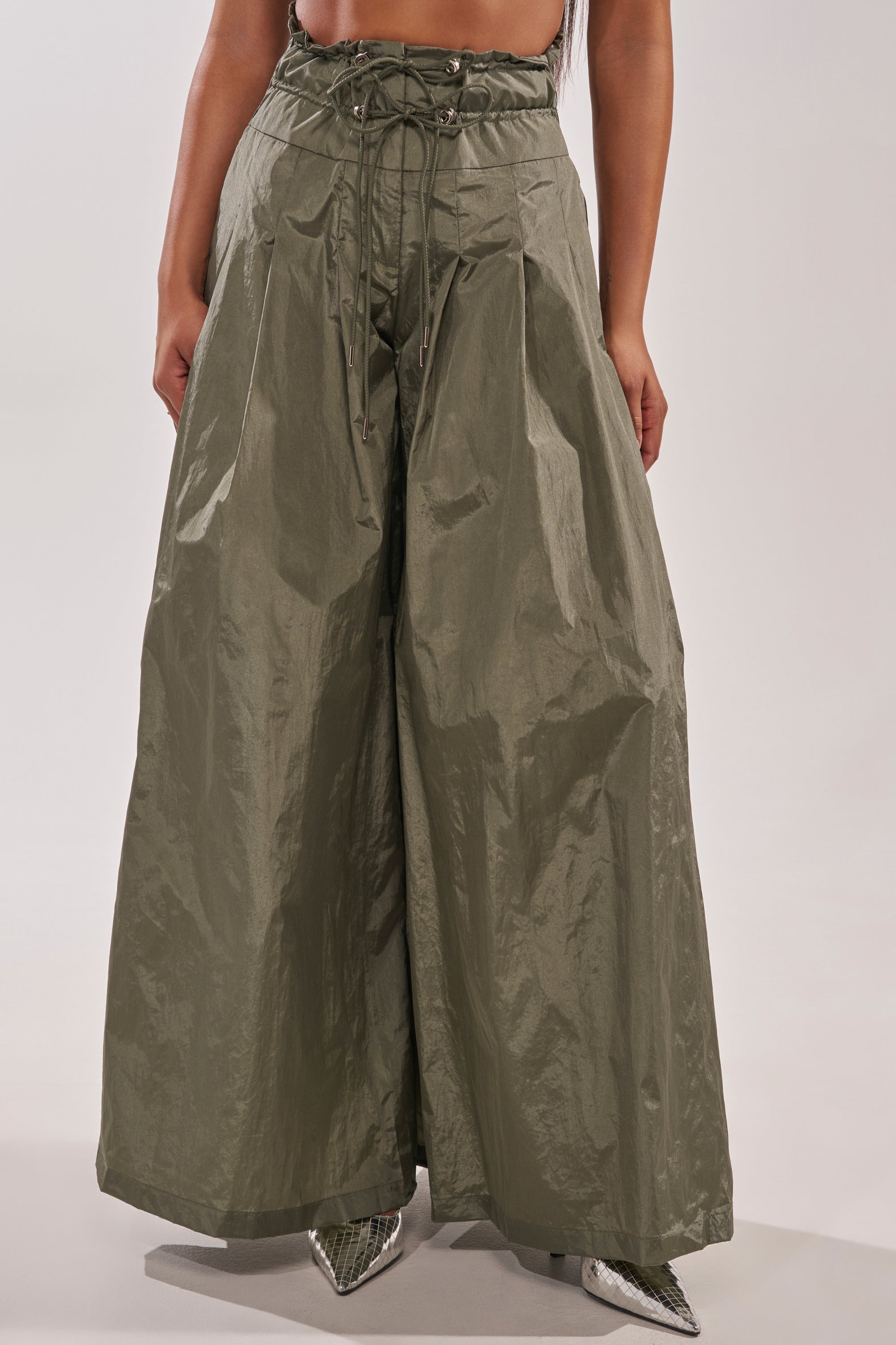 Someone in the DANCE THE NIGHT AWAY PALAZZO PANT—olive green, wide-leg, crinkled texture, lace-up high-rise waist—paired with pointed silver shoes. The upper body isn’t shown. 2