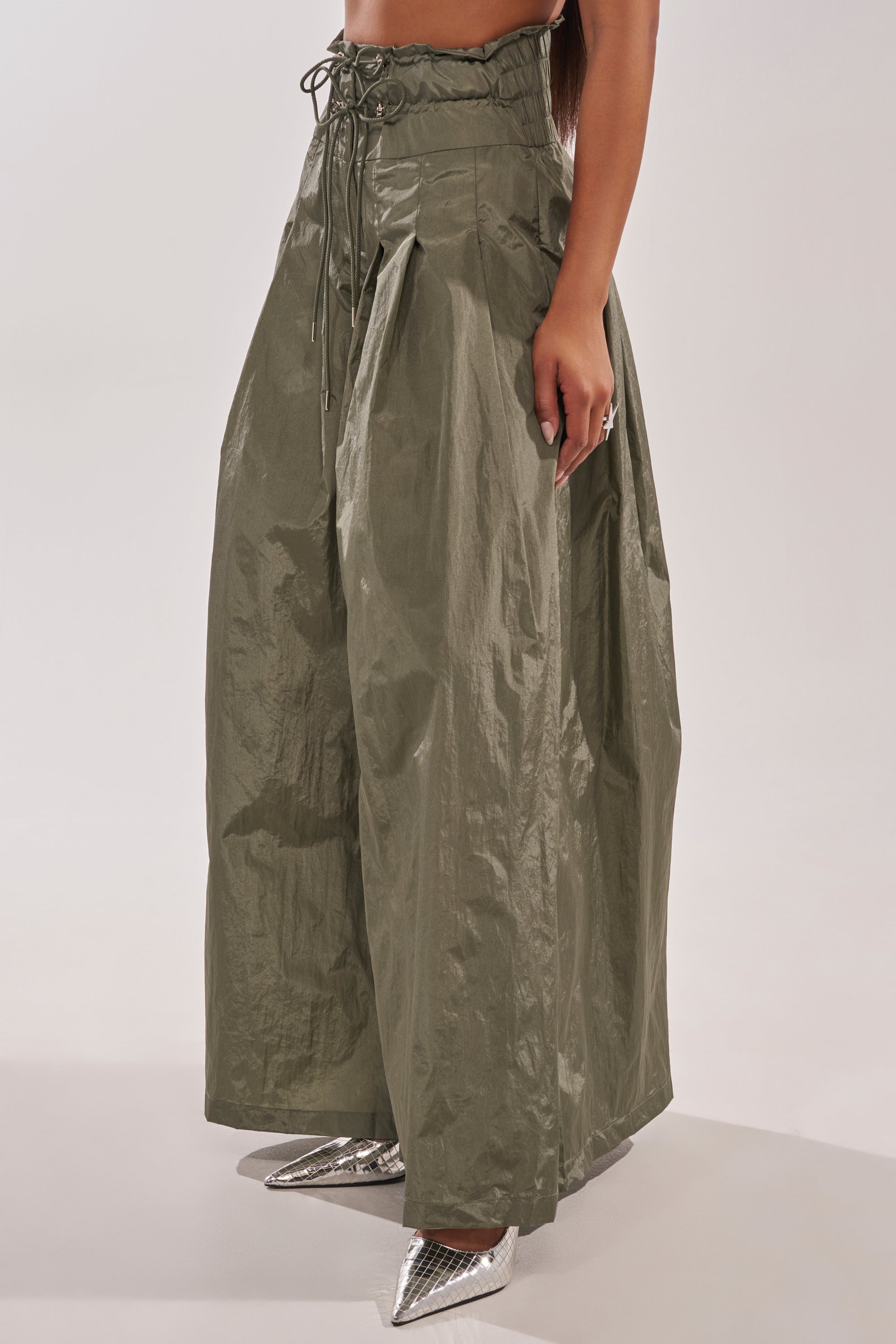 Someone stands against a plain background wearing DANCE THE NIGHT AWAY PALAZZO PANT, featuring high-rise, wide-leg utility style in olive green with a drawstring waist, paired with shiny silver pointed-toe shoes. 3