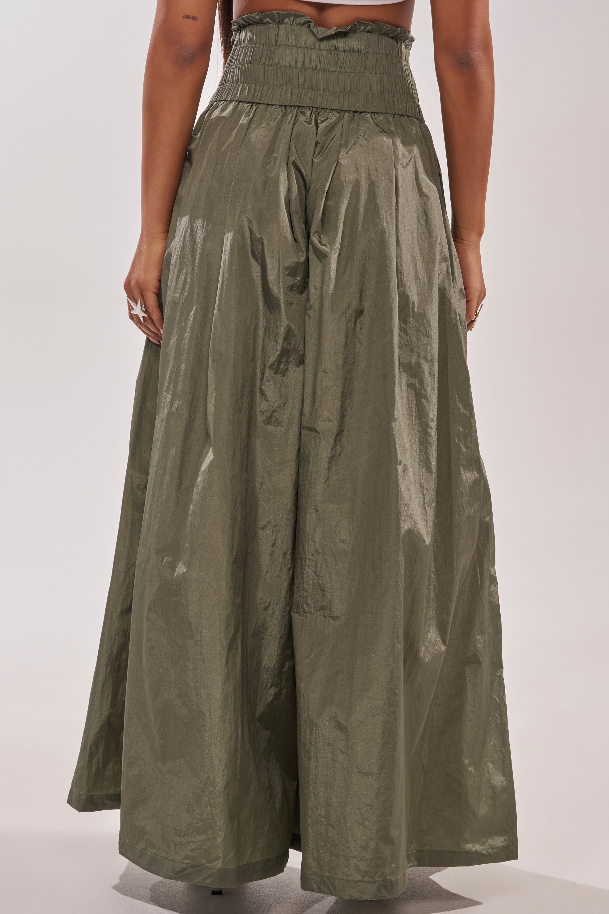 A person wears the DANCE THE NIGHT AWAY PALAZZO PANT, an olive green, high-rise, wide-leg pant made of shiny crinkled fabric, shown from the back against a plain background. 4