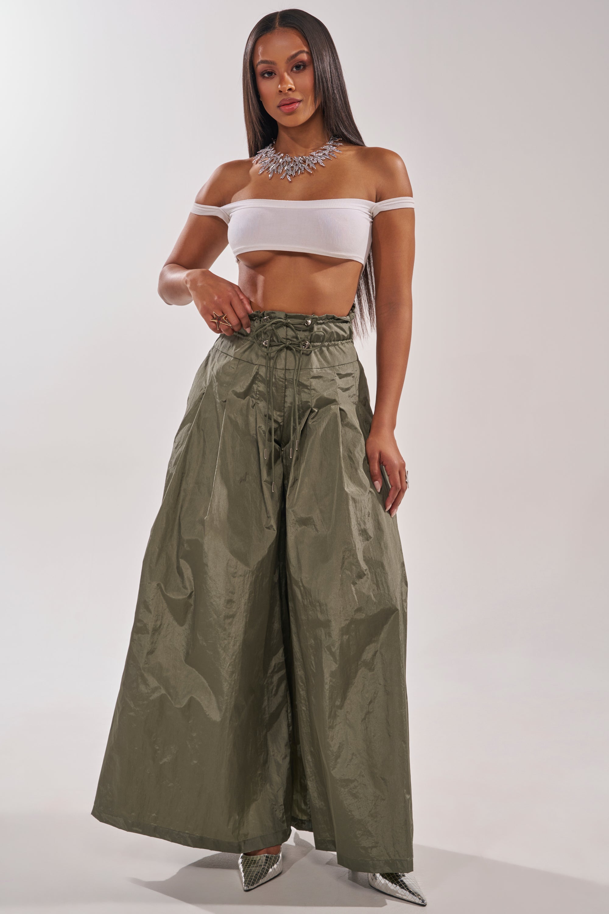 A woman poses in a sheer off-the-shoulder white crop top, DANCE THE NIGHT AWAY PALAZZO PANT in olive green with a high-rise drawstring waist, silver pointy heels, and a statement necklace on a plain background. 5