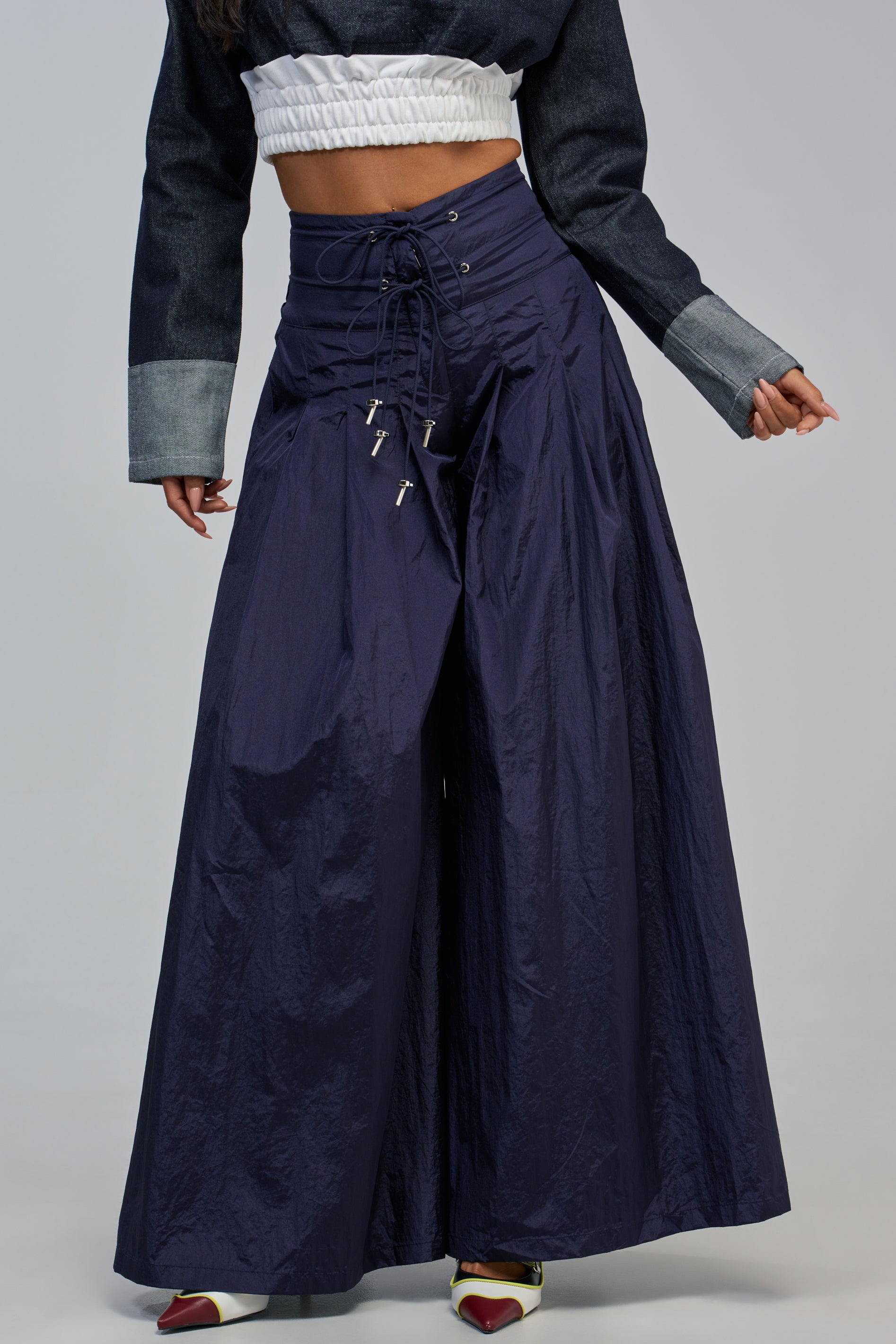 A person is seen wearing a cropped black and white jacket and the DANCE THE NIGHT AWAY PALAZZO PANT IN NAVY, which features a wide-leg silhouette with a lace-up front, styled with pointed white shoes featuring red tips. Only the body is visible, creating a sporty streetwear vibe.