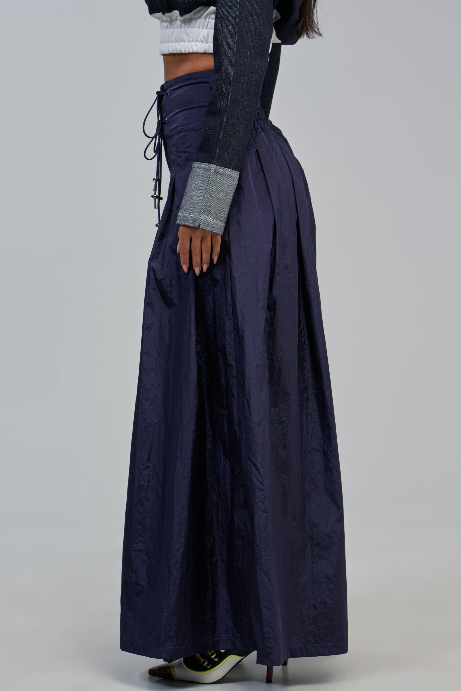A woman in the “Dance The Night Away Palazzo Pant in Navy,” featuring a long, wide-leg silhouette with a gathered waistband and lace-up back, pairs the look with a cropped black and gray jacket as she stands sideways against a plain light background, creating an elegant yet subtly sporty streetwear vibe.